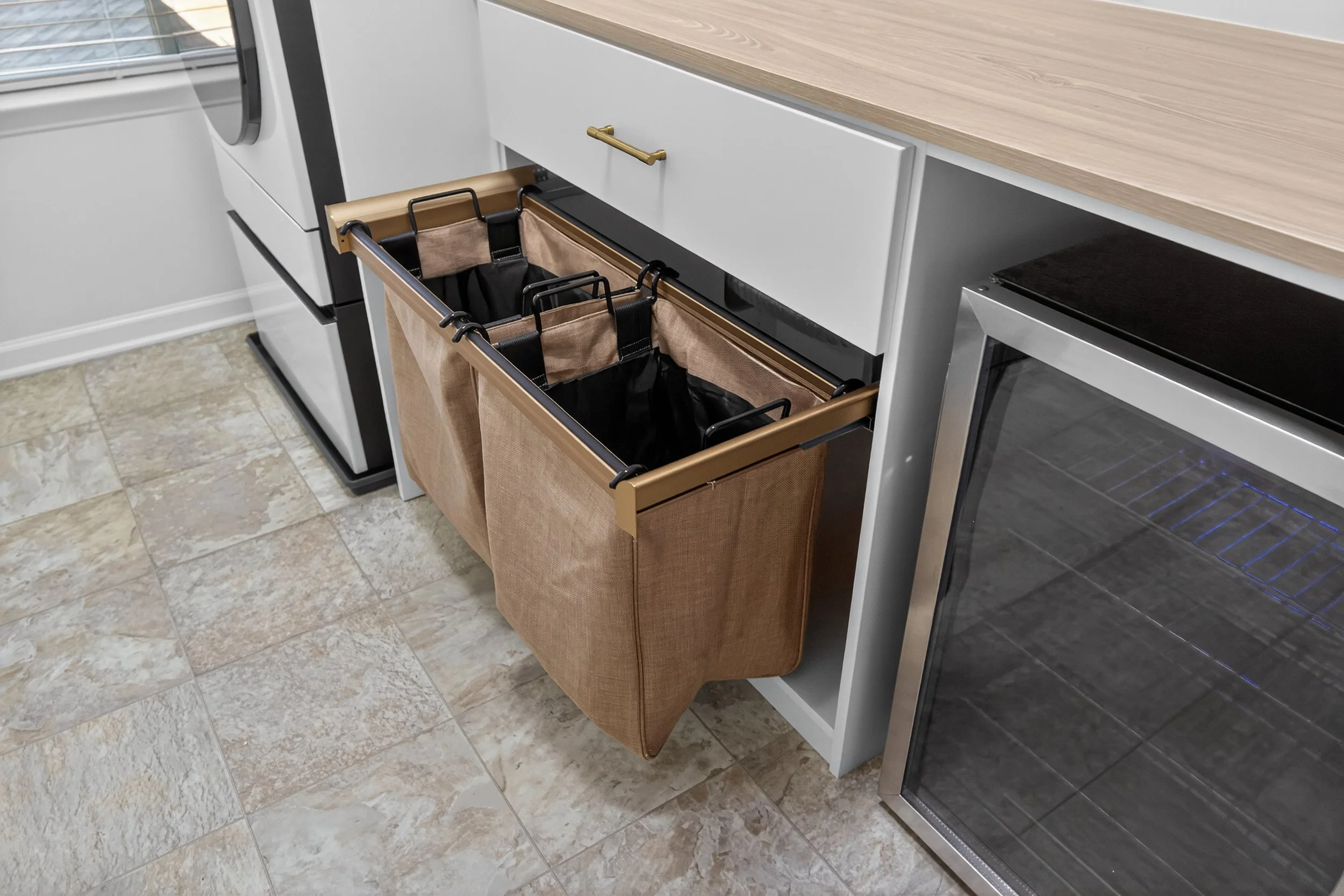 Custom pull-out TAG laundry hamper with soft-close hardware in a luxury laundry room, paired with a built-in beverage fridge for effortless organization and everyday comfort.