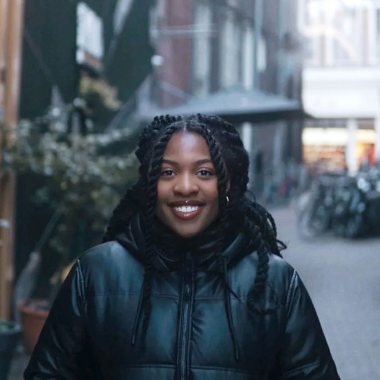 A young woman with dark curly hair in twists smiling outdoors in an urban setting, wearing a black leather jacket.