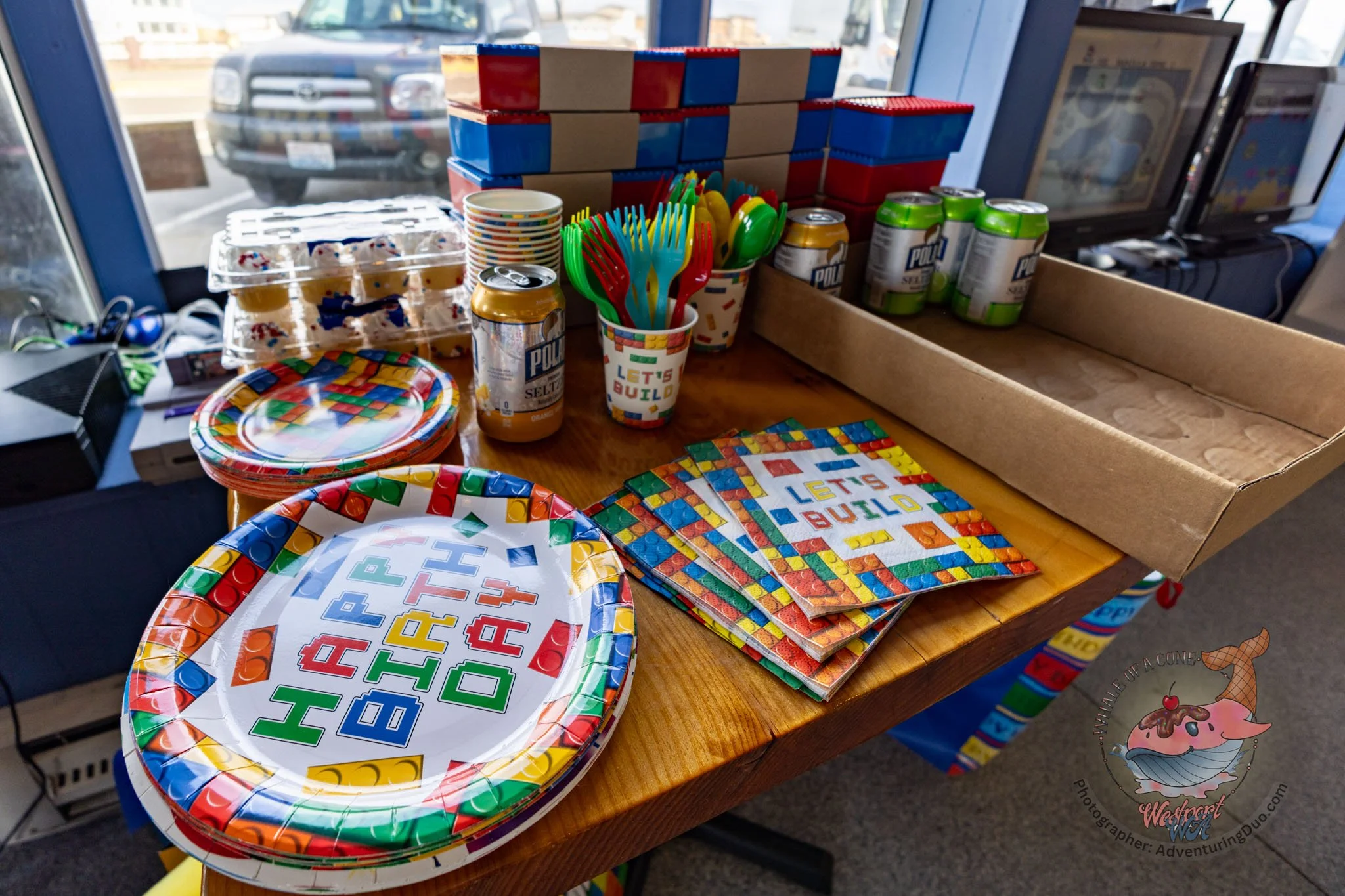 A table with party supplies including colorful paper plates, cups, napkins with alphabet and number designs, plastic utensils in a cup with the phrase 'Let's Build', cans of sparkling water, and a cardboard box for organizing or serving. In the backg