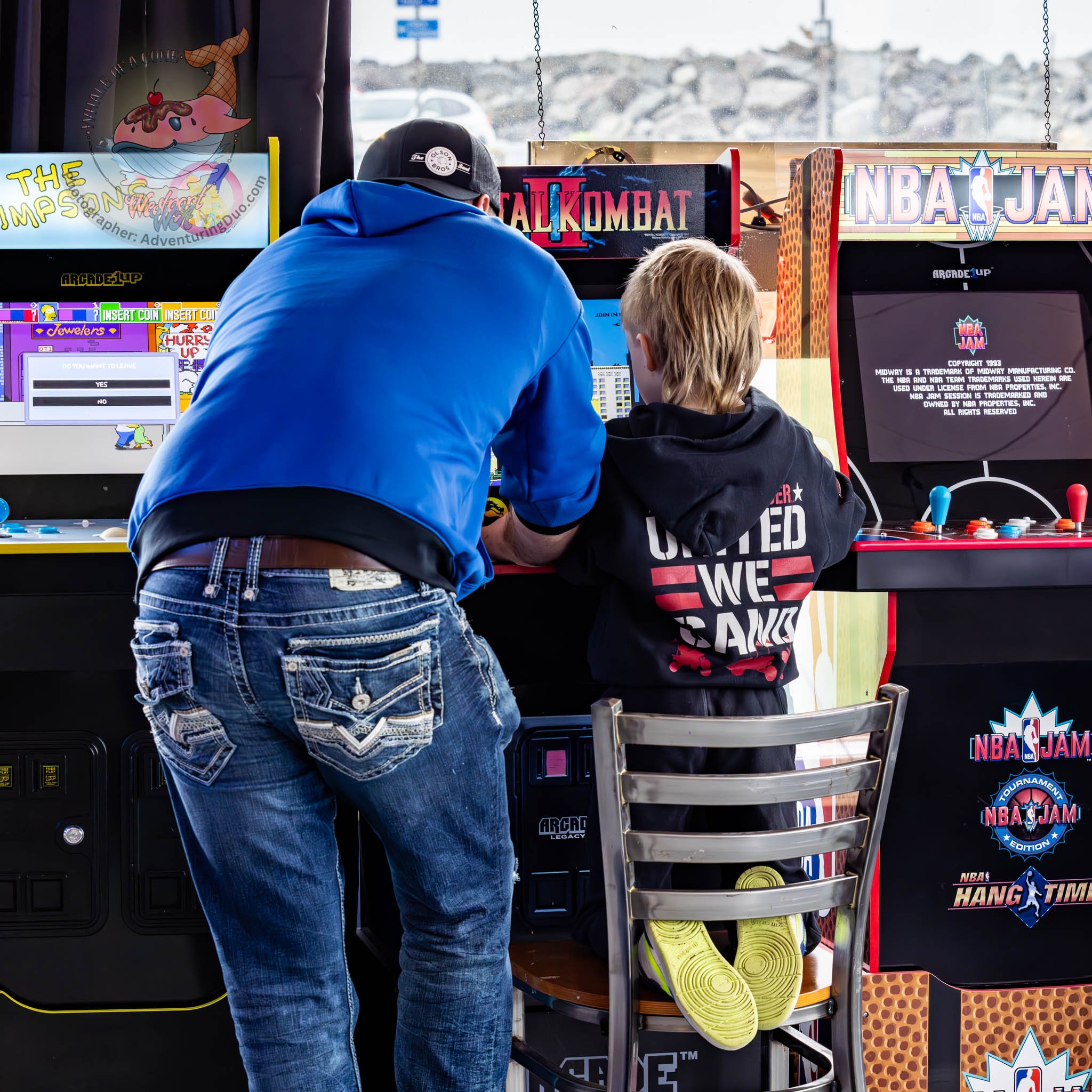 Two people, possibly a man and a boy, sitting at a row of arcade racing games inside an arcade or gaming center. The man is wearing a black cap and a blue hoodie, while the boy is dressed in black with a printed jacket. The arcade games are themed ar