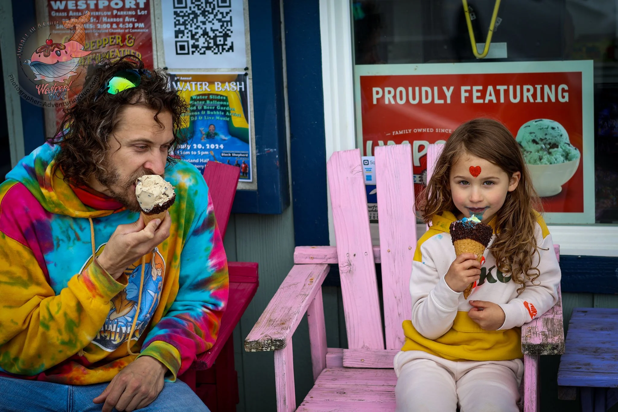 A man and a girl sitting on pink and purple benches outside an ice cream shop, enjoying ice cream cones. The man wears a colorful tie-dye hoodie and the girl has a red heart sticker on her forehead.