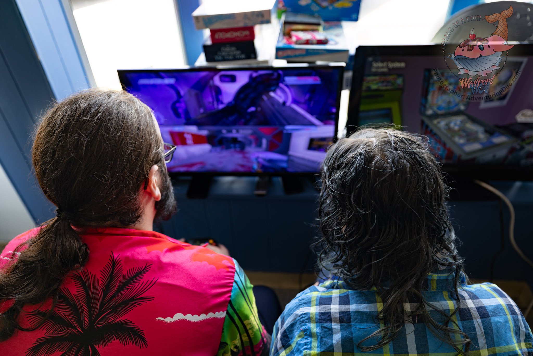 Two children with long hair, sitting outdoors and playing video games on a computer with purple lighting. One child is wearing a bright pink shirt with black palm trees, and the other is wearing a blue plaid shirt.