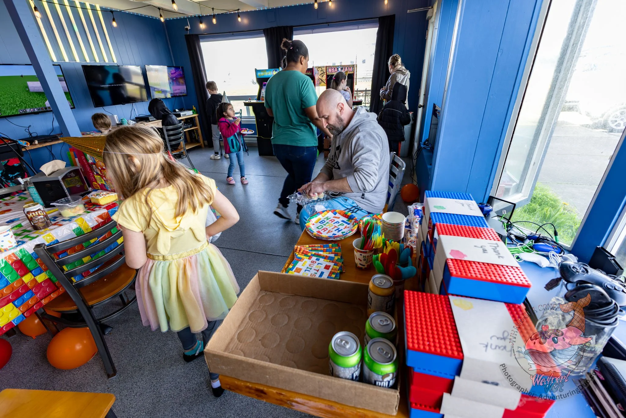 Children and adults at a colorful birthday party with games, balloons, and a cake in a blue room decorated with string lights.