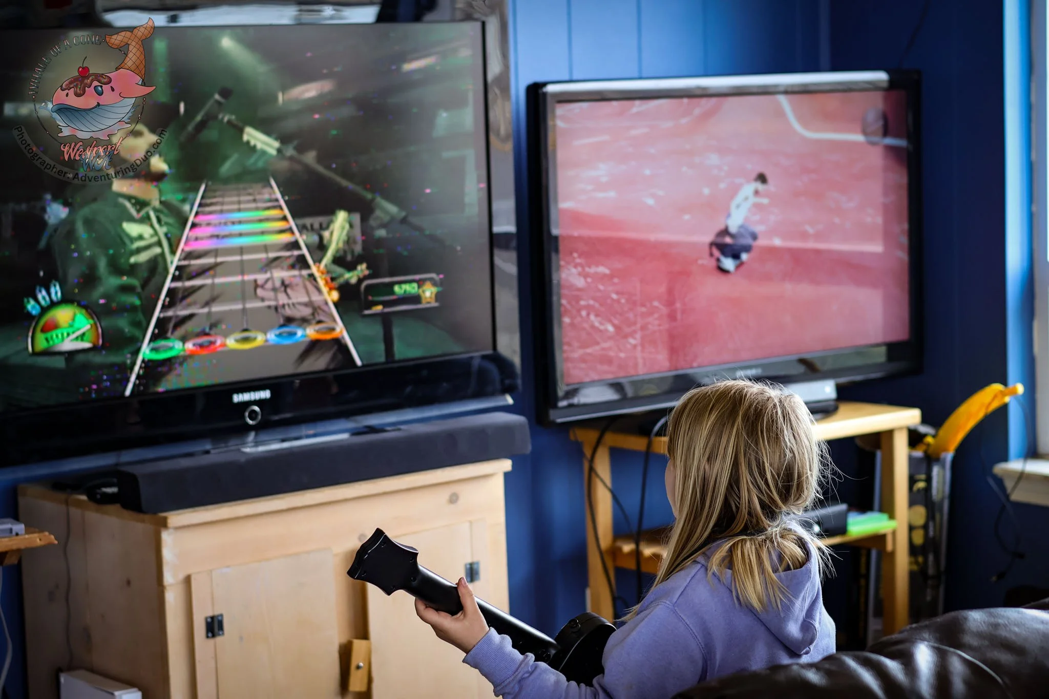 A young girl with blonde hair is playing a video game on a large screen, holding a gaming controller, seated in a room with wooden furniture and blue walls.