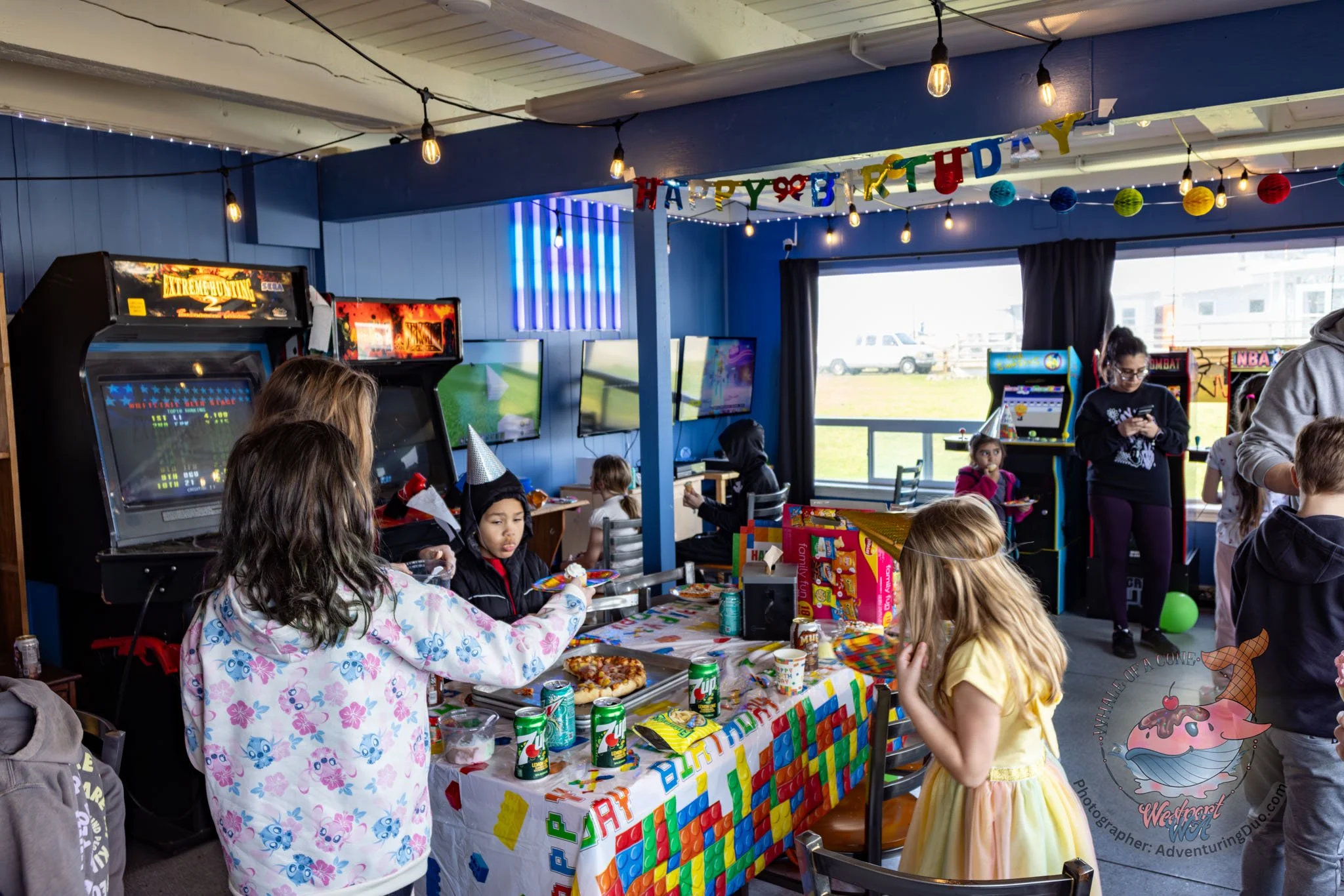 Children at a birthday party with a 'Happy Birthday' banner, arcade games, and a table with pizza, snacks, and drinks.