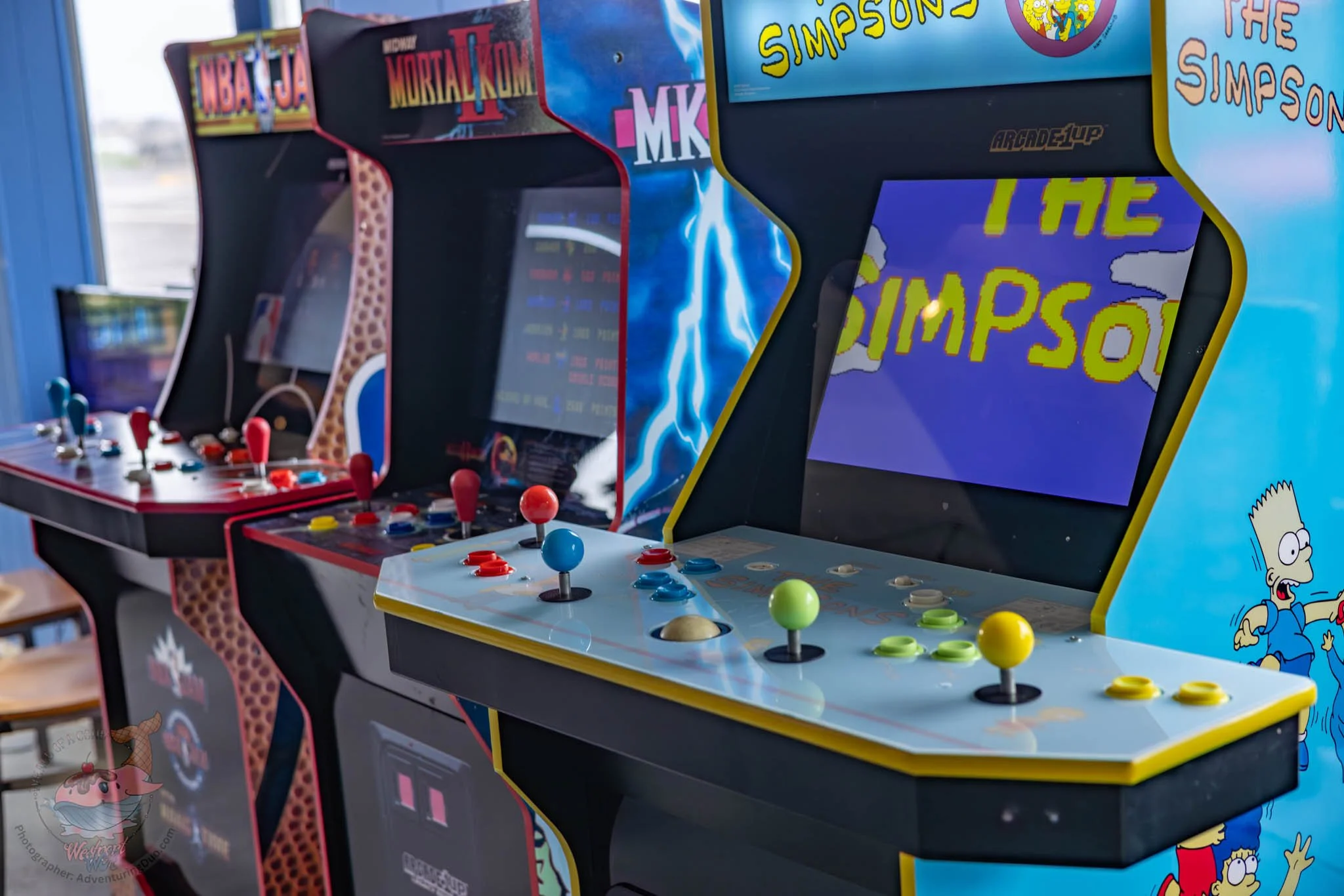 Several vintage arcade machines are lined up, featuring popular characters and themes from classic video games and TV shows, including The Simpsons, Mortal Kombat, and NBA Jams.