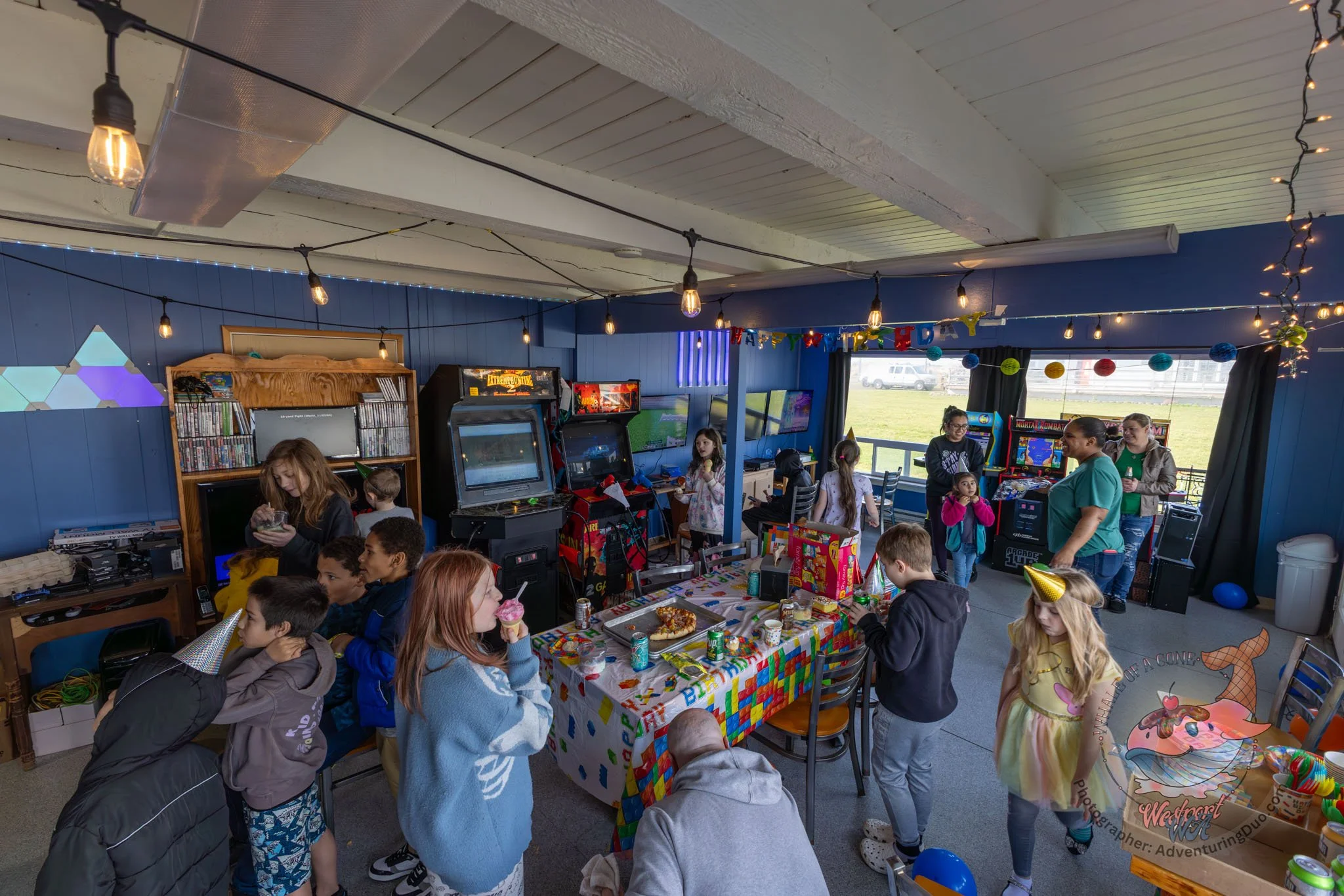 Children and adults celebrating at a birthday party indoors with decorations, snacks, and arcade games.