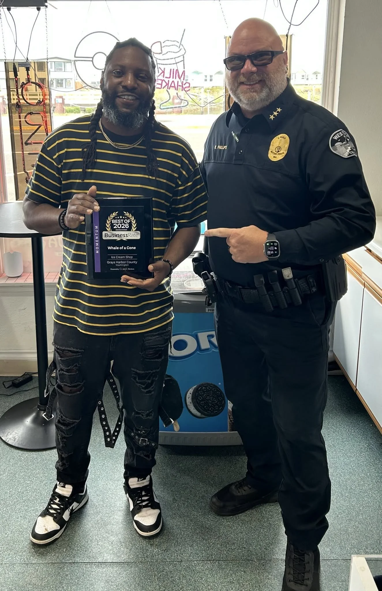 A man with braided hair and a beard holding a plaque stands next to a police officer smiling in an office or store setting. The plaque reads 'Best of 2023' and 'Whale of a Cone' in Grays Harbor County. The police officer wears a uniform with a badge and a patch on the sleeve, and is pointing at the man. The background includes a window, a neon sign, and some equipment.