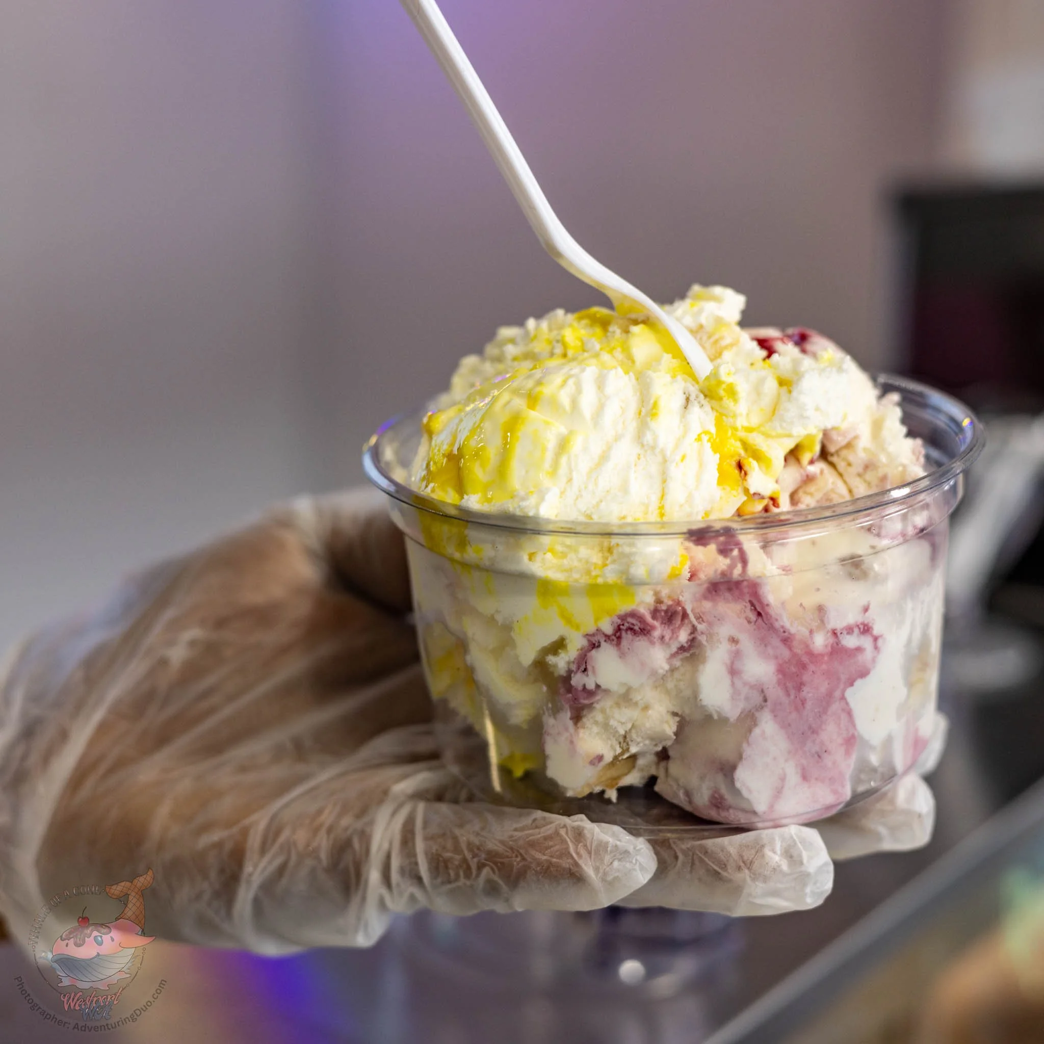 Close-up of a bowl of ice cream topped with yellow sauce and chunks, with a plastic spoon inserted, placed on a surface.