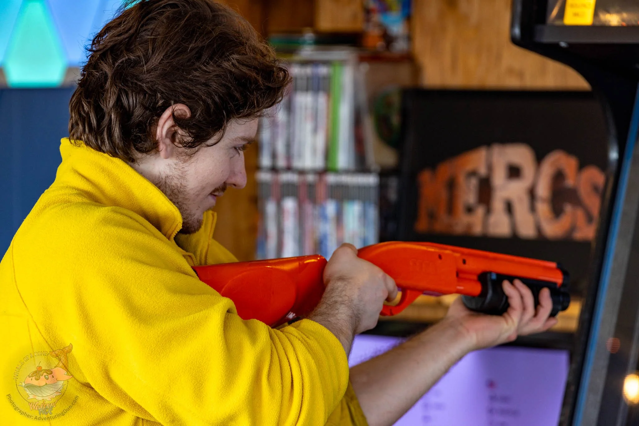 A young man with curly brown hair wearing a yellow hoodie aiming an orange toy gun in an indoor setting.