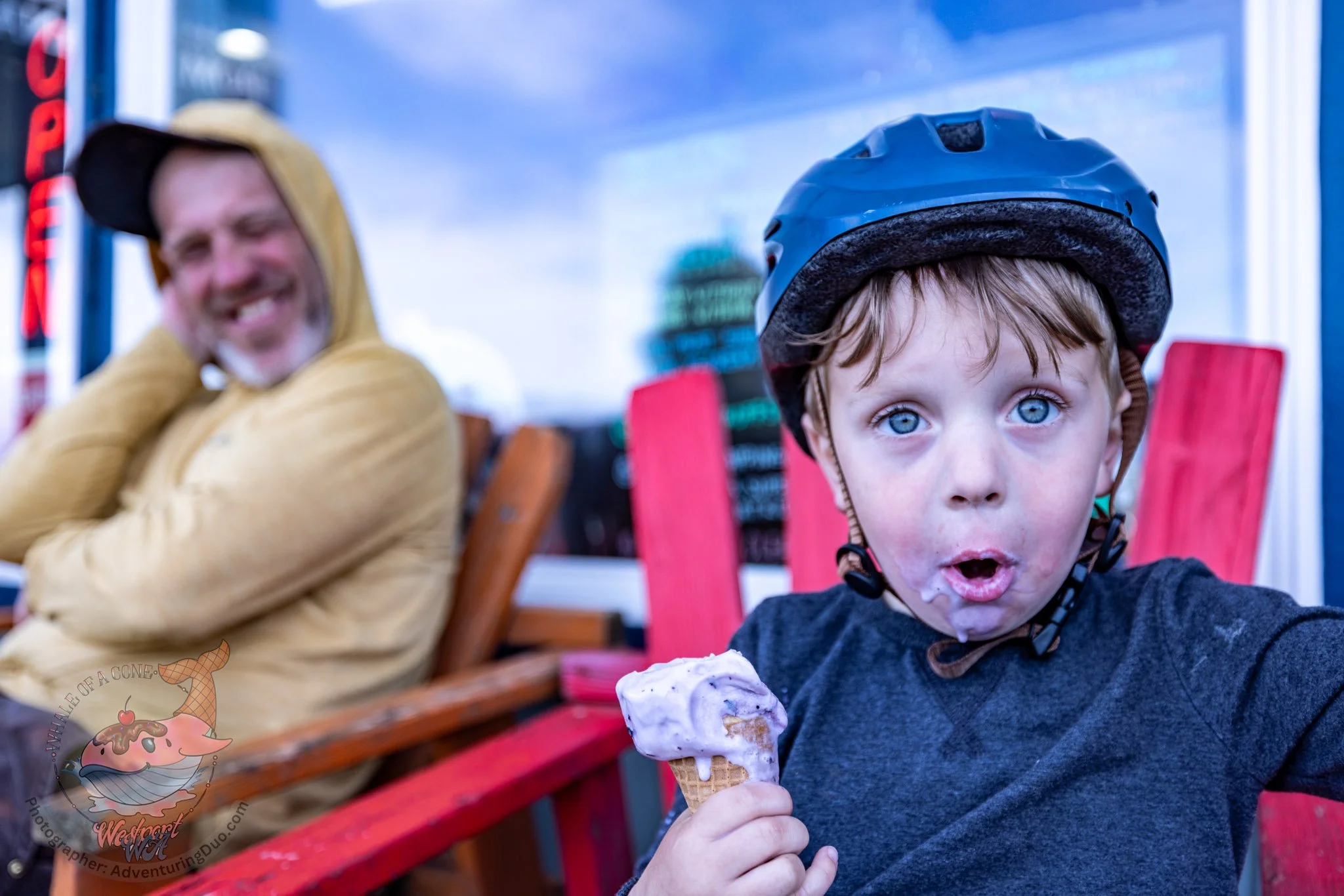 A young boy with blue eyes and a bike helmet is holding a melting ice cream cone with pink and white ice cream, sitting on a red bench with an adult man smiling in the background.