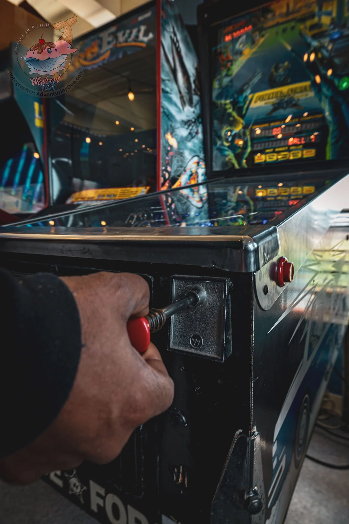 Close-up of a person's hand pressing the button on a pinball machine in a gaming arcade.