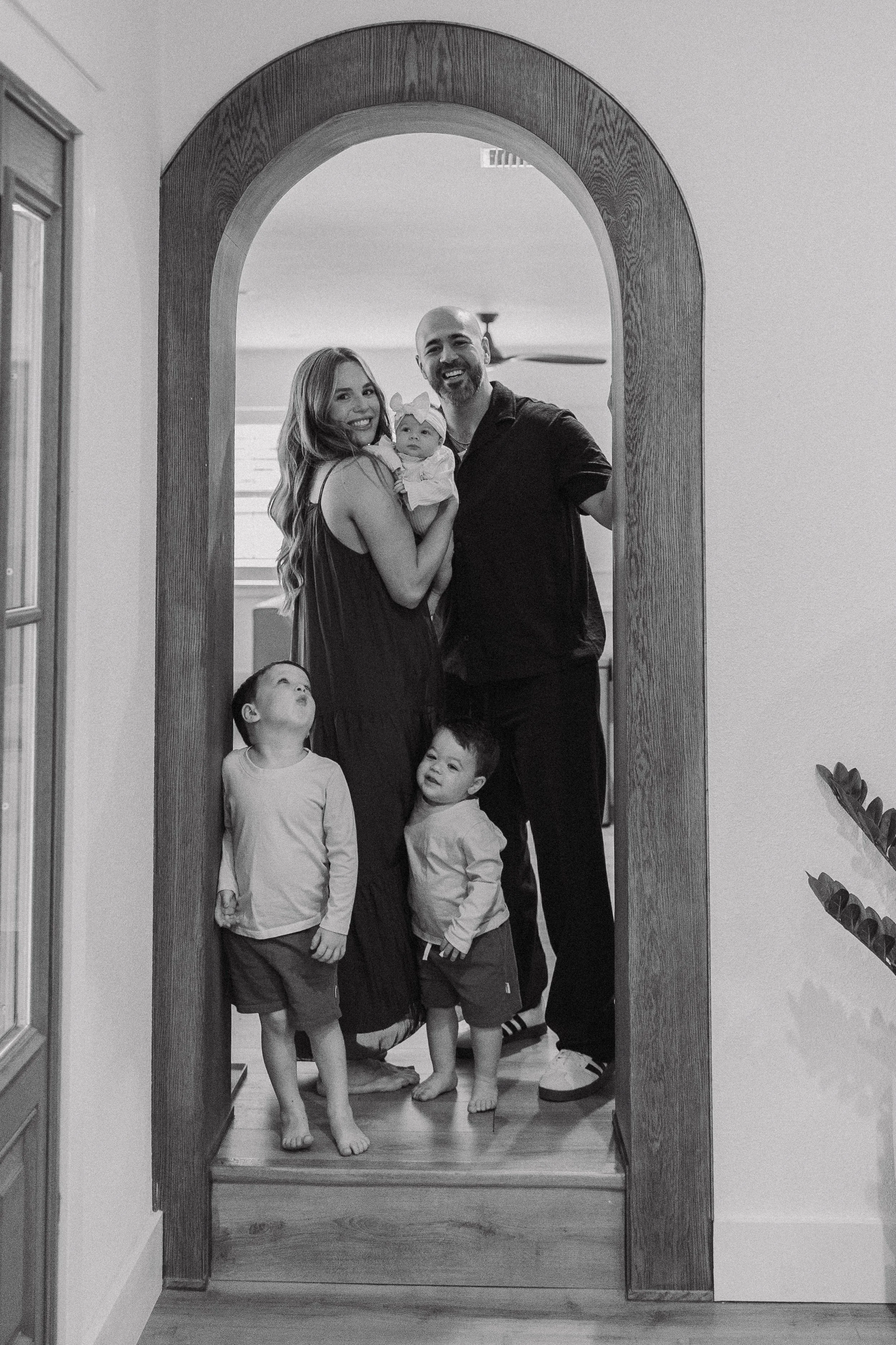 A black and white photo of a family of five standing in a doorway. The woman is holding a baby girl, and two young boys are standing in front of them. They are smiling and looking cheerful.