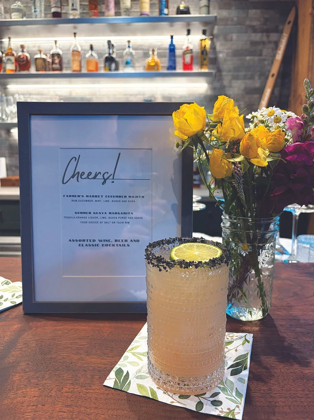 A cocktail with a slice of lime garnish on top, placed on a floral napkin on a wooden table, with a sign listing drinks and a bouquet of yellow, purple, and white flowers in a glass jar in the background.