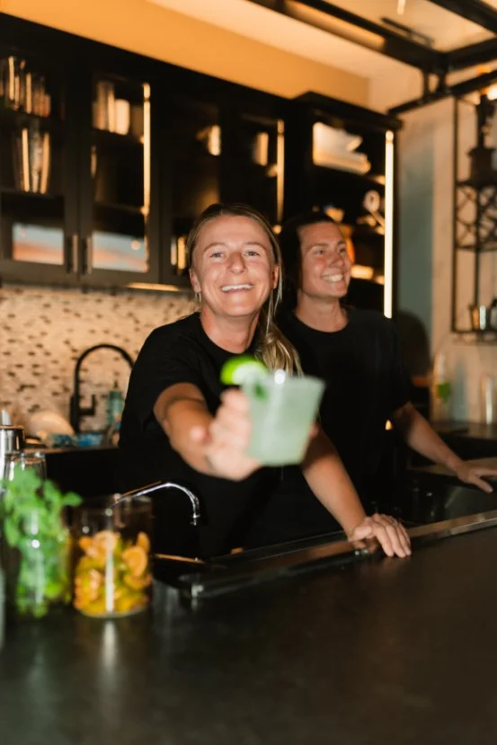smiling bartenders handing you a light green cocktails