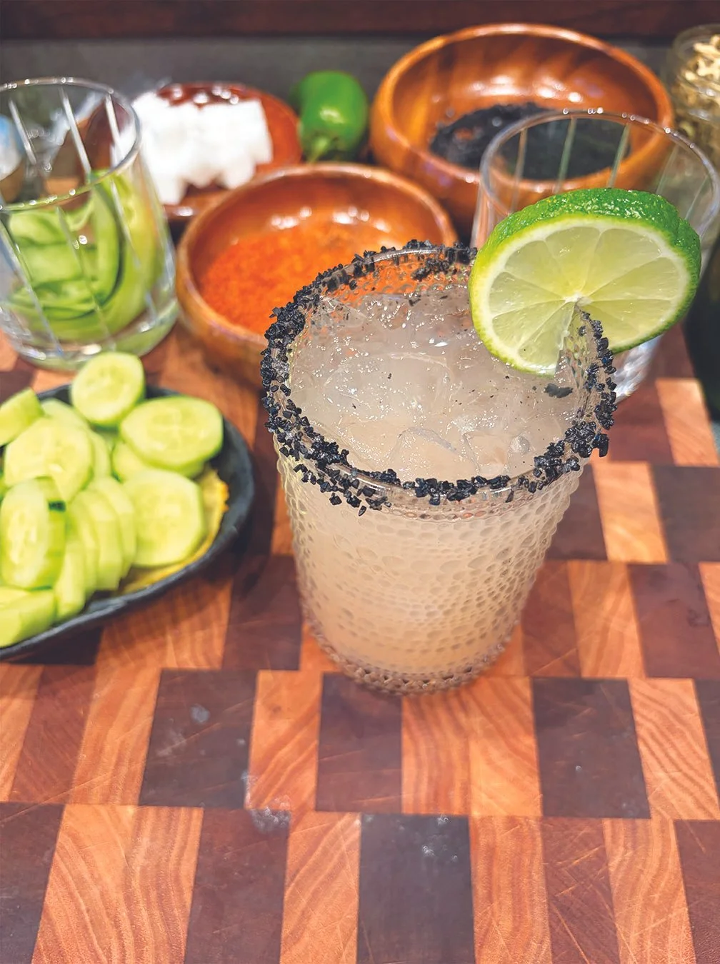 A margarita with a salted rim and lime wedge on top, placed on a wooden table with garnishes including sliced cucumbers, sliced green peppers, salt, Tajin, and other spices in small bowls.