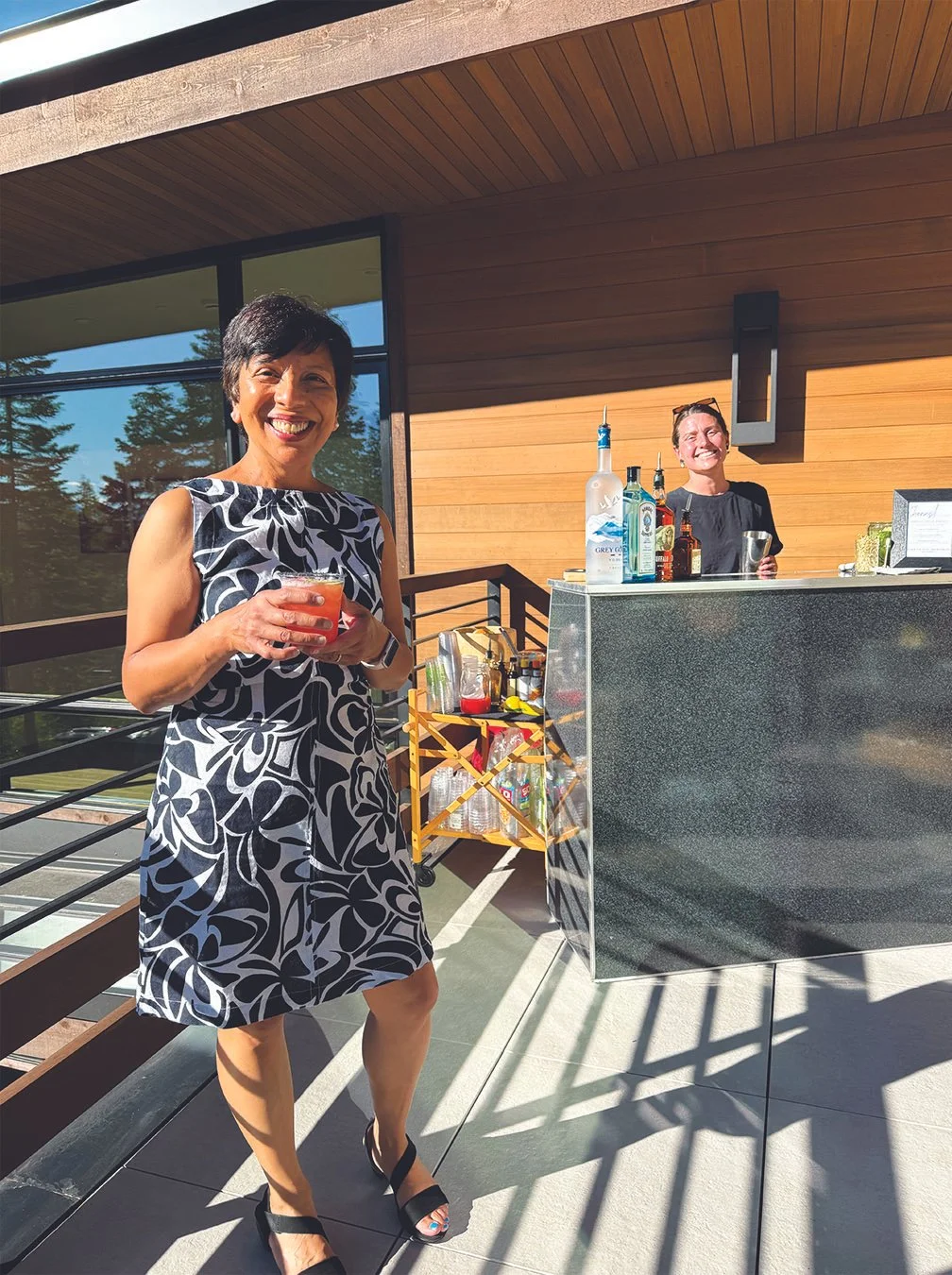 A woman in a black and white patterned dress holding a drink, standing outdoors on a balcony near a bar with another woman behind it, during daytime.
