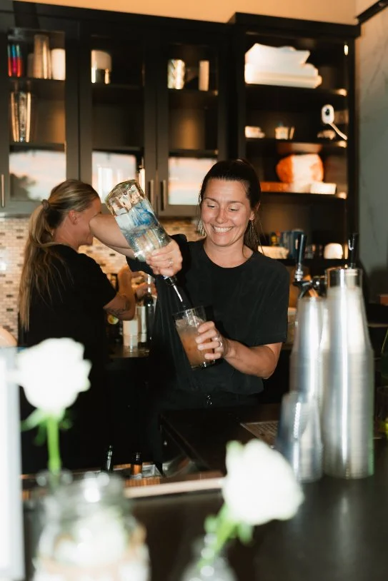 A smiling female bartender in a black shirt is pouring a drink from a large bottle into a glass in a bar or restaurant setting, with another woman in the background working behind the counter and shelves with bottles and kitchen items on the wall.