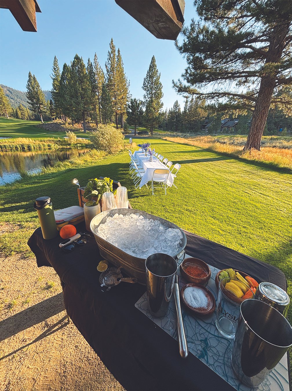 Outdoor dining setup by a lake with a long table and white chairs, surrounded by trees and open grassy area, with a table in the foreground holding ice, drinks, and snacks.