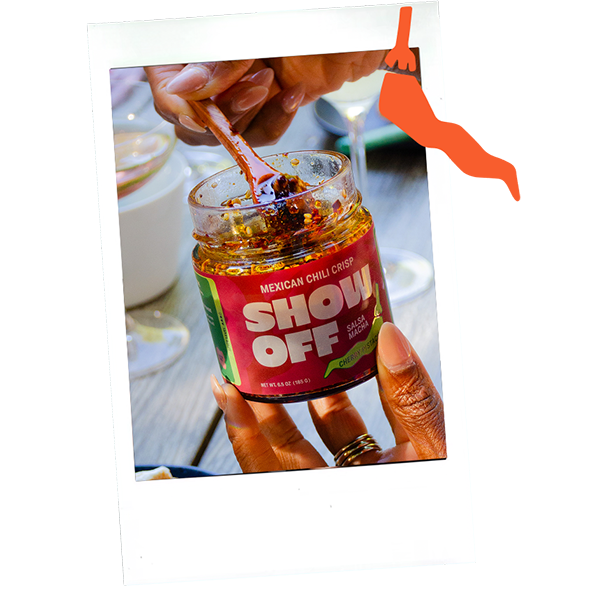 Hand holding a jar of Show Off Mexican chili salsa with a spoon inside, showing some of the salsa. The jar label is red and green with bold white text.