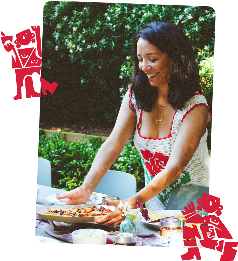 A woman setting a plate of food at an outdoor table with lush green bushes in the background.