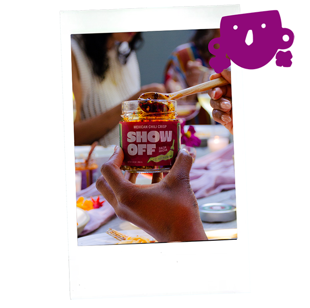 Close-up of hand holding a jar of Show Off Mexican chili crisp with a wooden spoon on top, at a table with people in the background.