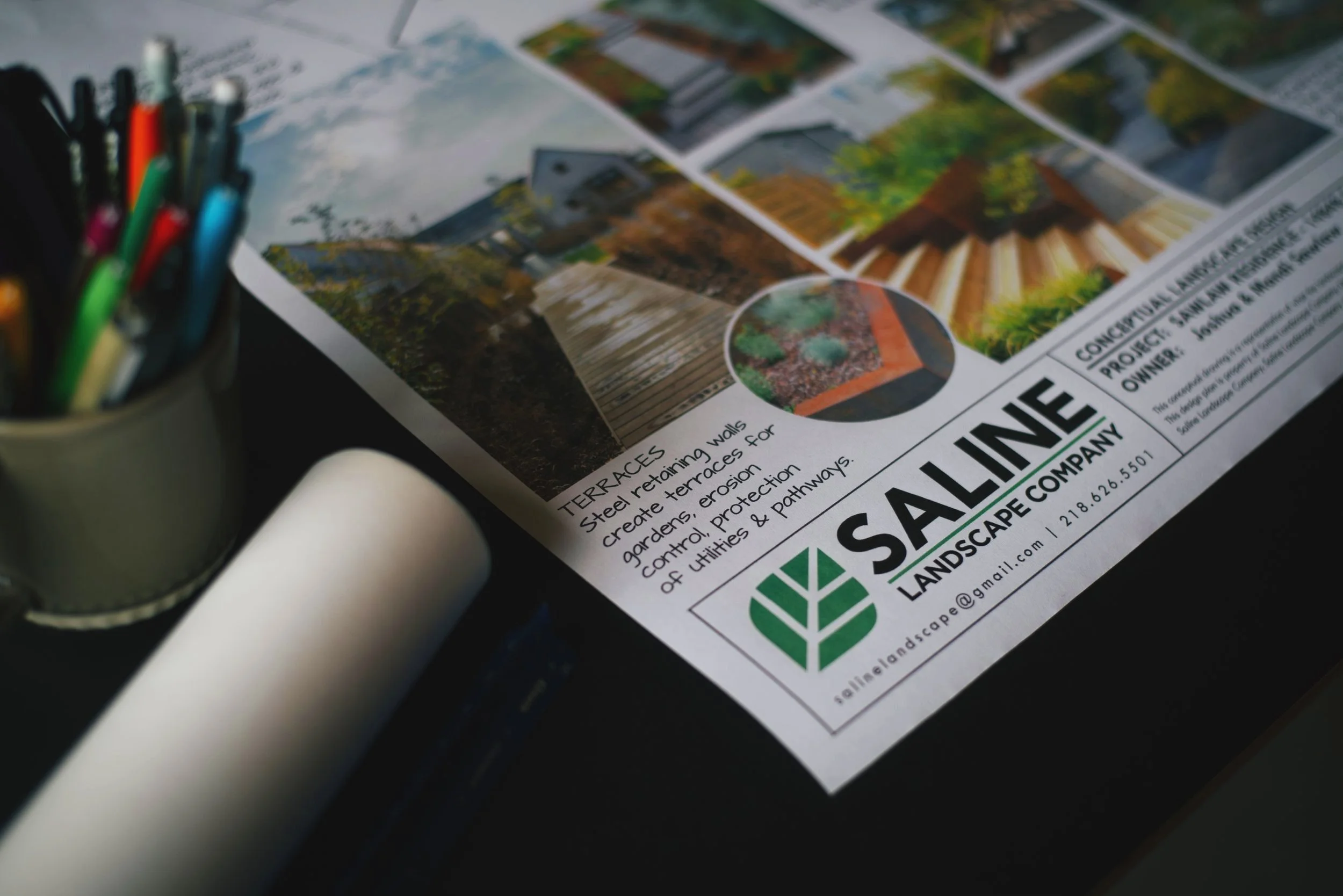A landscape design company's brochure showing images of gardens, pathways, and walls, with a pen holder, marker, and rolled-up paper on a dark surface.