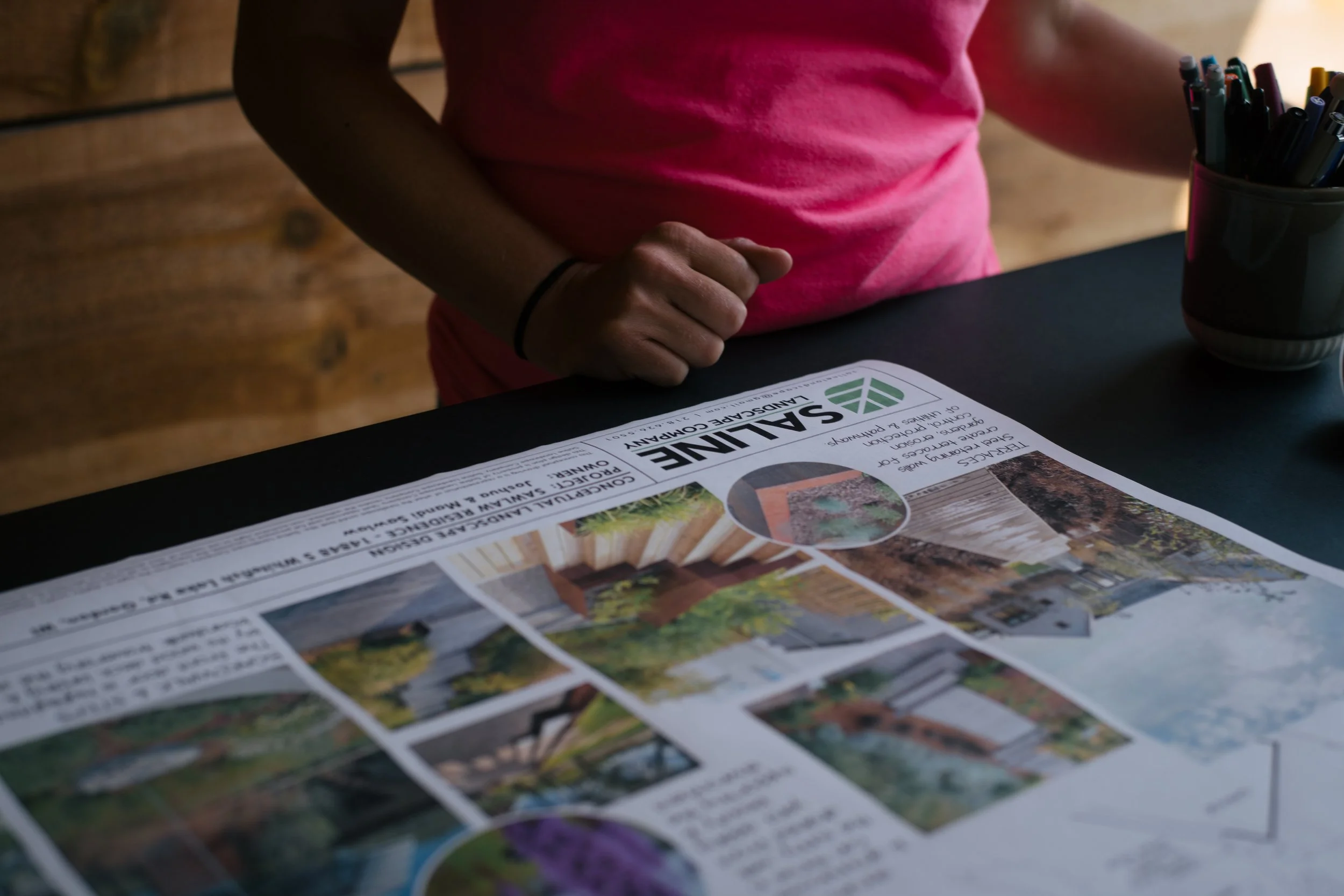 Person in pink shirt reading a landscaping newsletter with images of outdoor spaces, sitting at a black table with a container of pens.