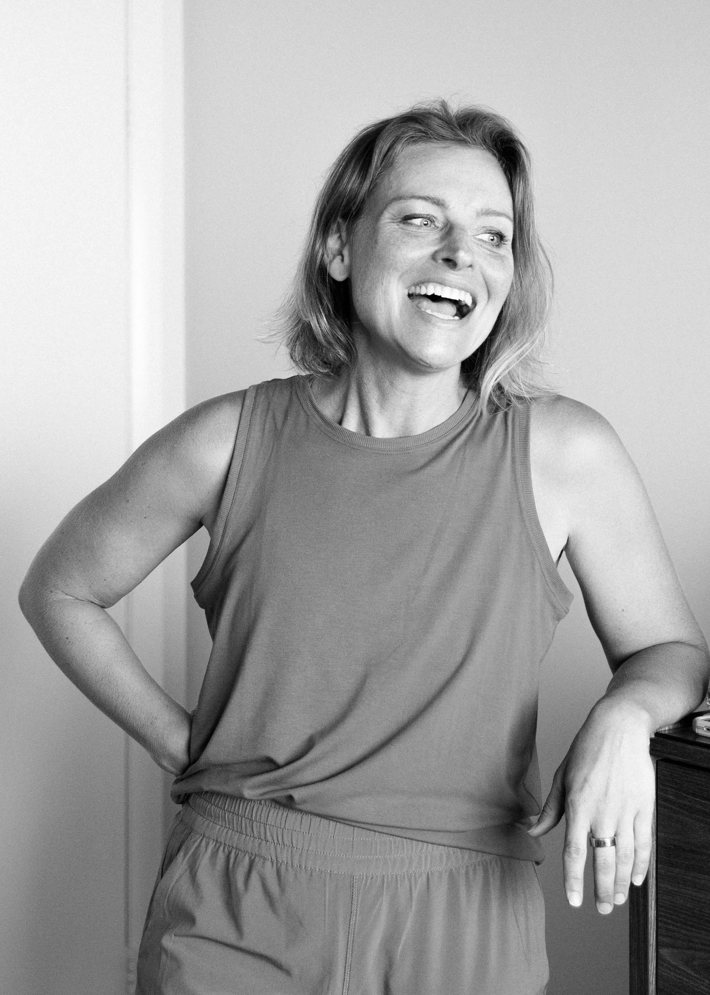 Black and white photo of Victoria Steward, laughing and wearing a sleeveless top, leaning casually against a piece of furniture.