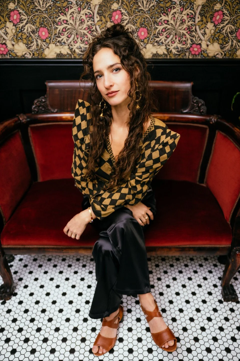 Fabienne Annick seated on a red velvet vintage sofa with patterned wallpaper behind her, wearing a checkered gold and black top, satin pants, and statement earrings, looking calmly at the camera with a poised expression.