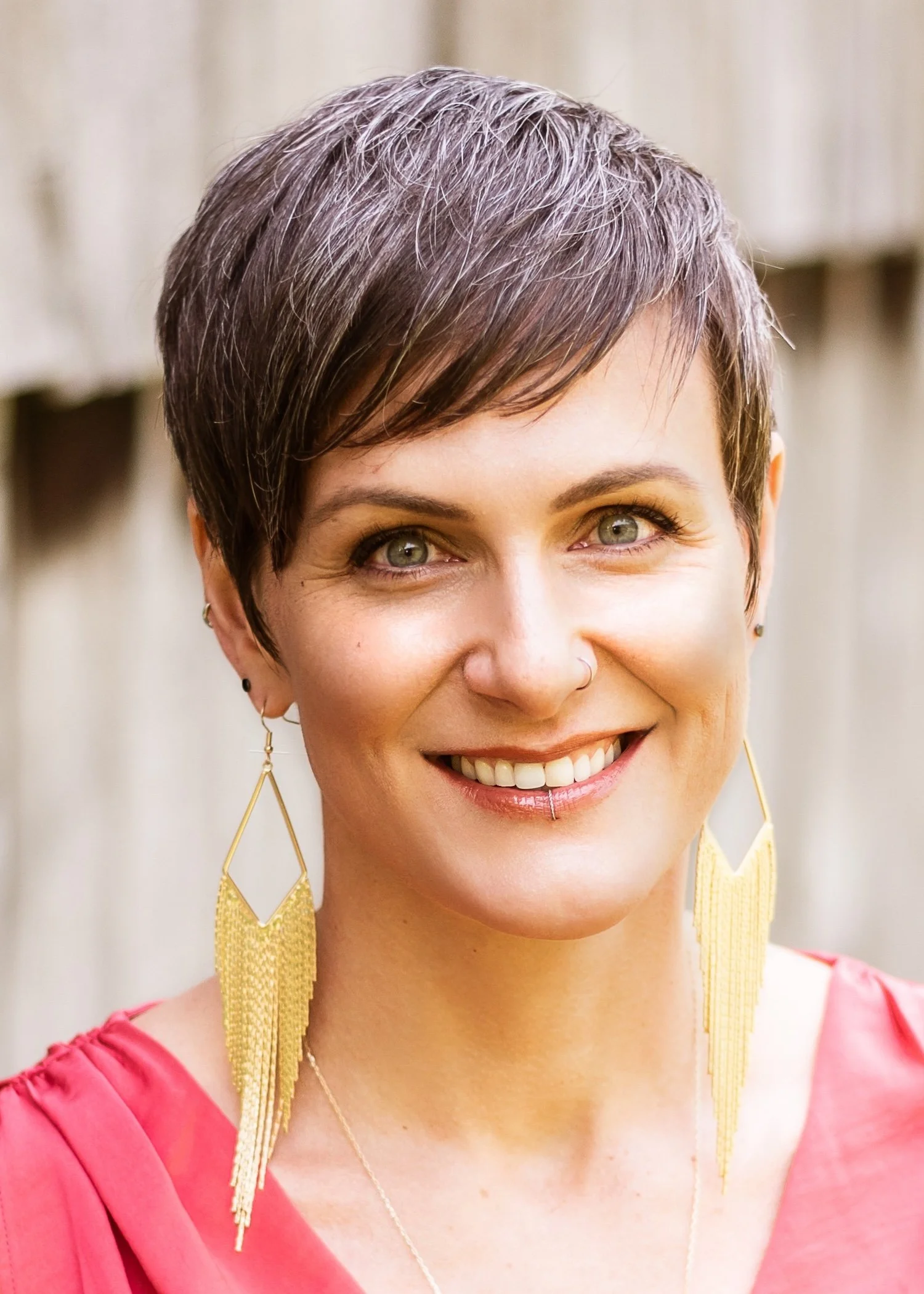 Cory Scott smiling in soft natural light, wearing a coral top and bold gold fringe earrings, with short salt-and-pepper hair and a radiant, confident presence.