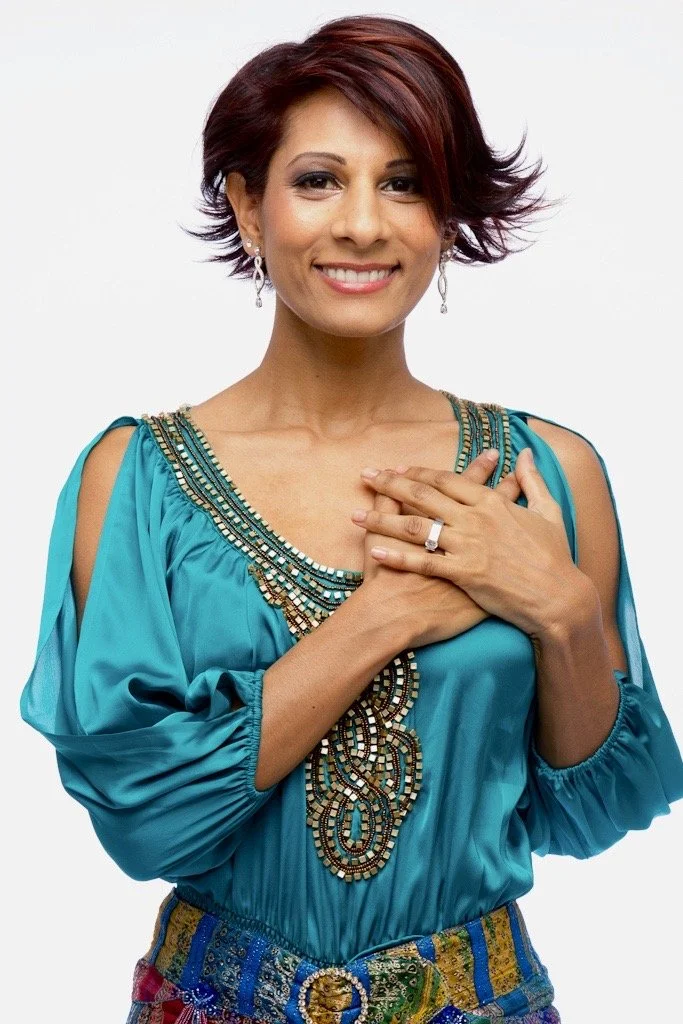 Annie Lalla smiling softly with both hands over her heart, wearing a teal top with intricate beading and colorful embroidered belt, standing against a white background with radiant presence.