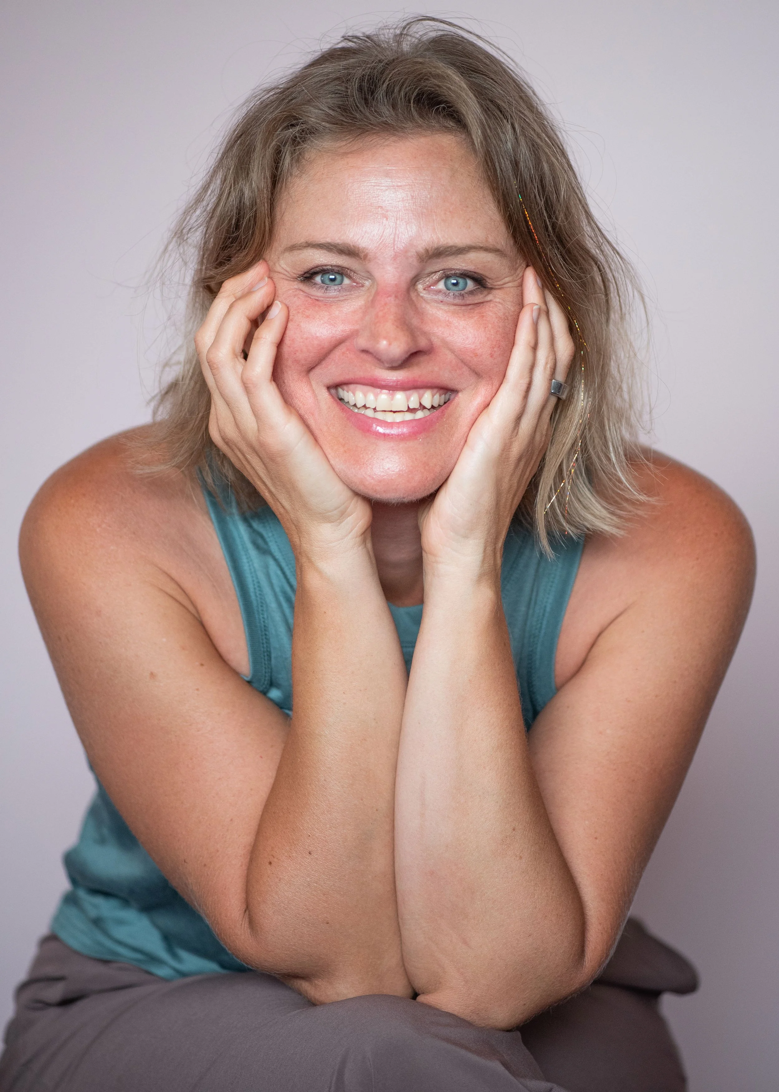Victoria Steward smiling warmly with her hands resting on her cheeks, wearing a teal tank top and seated comfortably, radiating grounded joy and approachability.