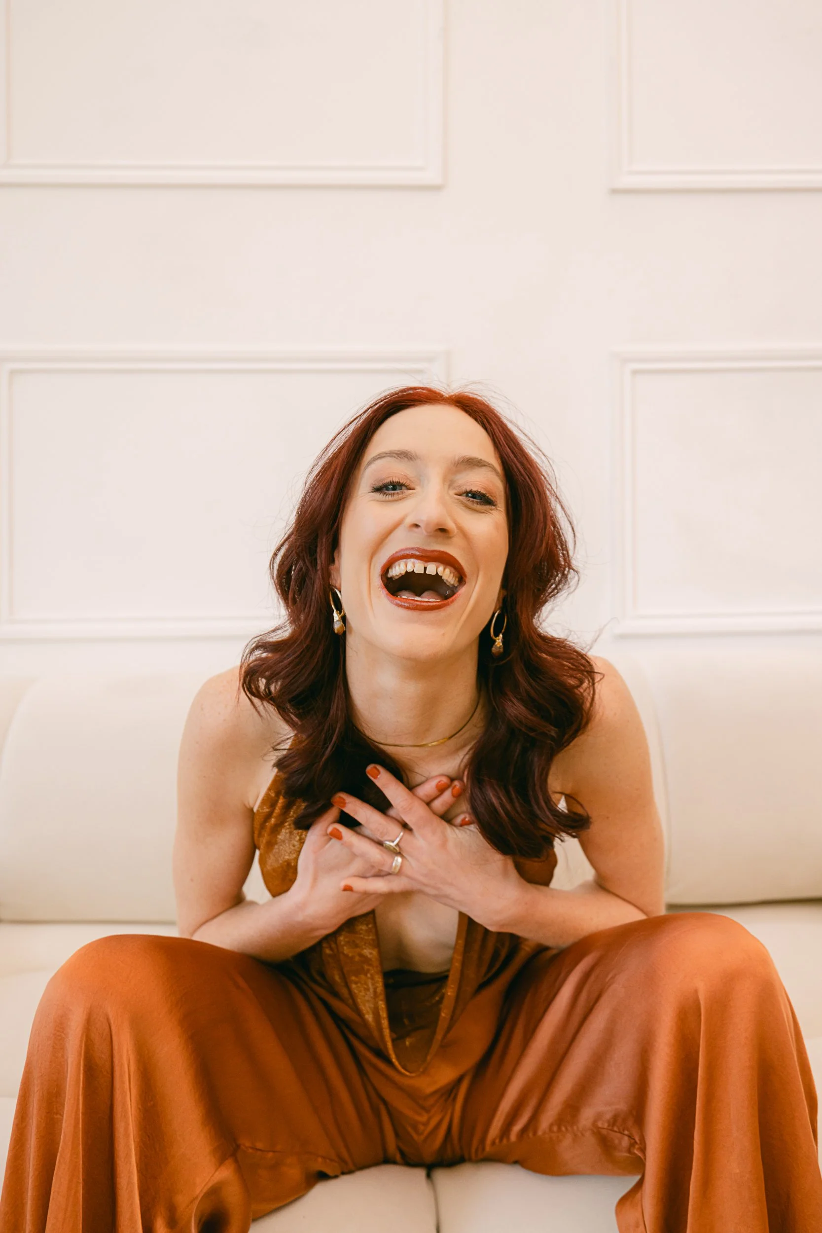Rosa Maxwell laughing joyfully while seated on a light-colored couch, wearing a rust-colored jumpsuit and gold jewelry, hands over heart in a playful, expressive pose.