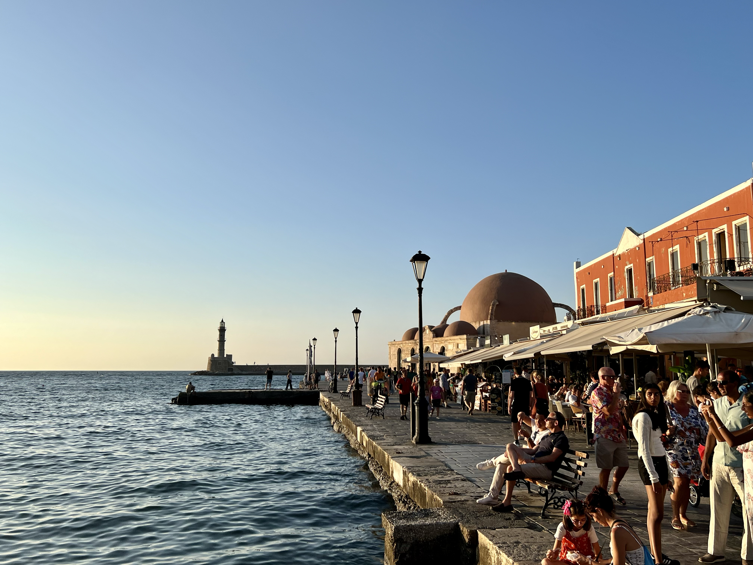 People walking and sitting along a seaside promenade at sunset with a lighthouse in the distance, outdoor cafe with awnings, and colorful buildings.