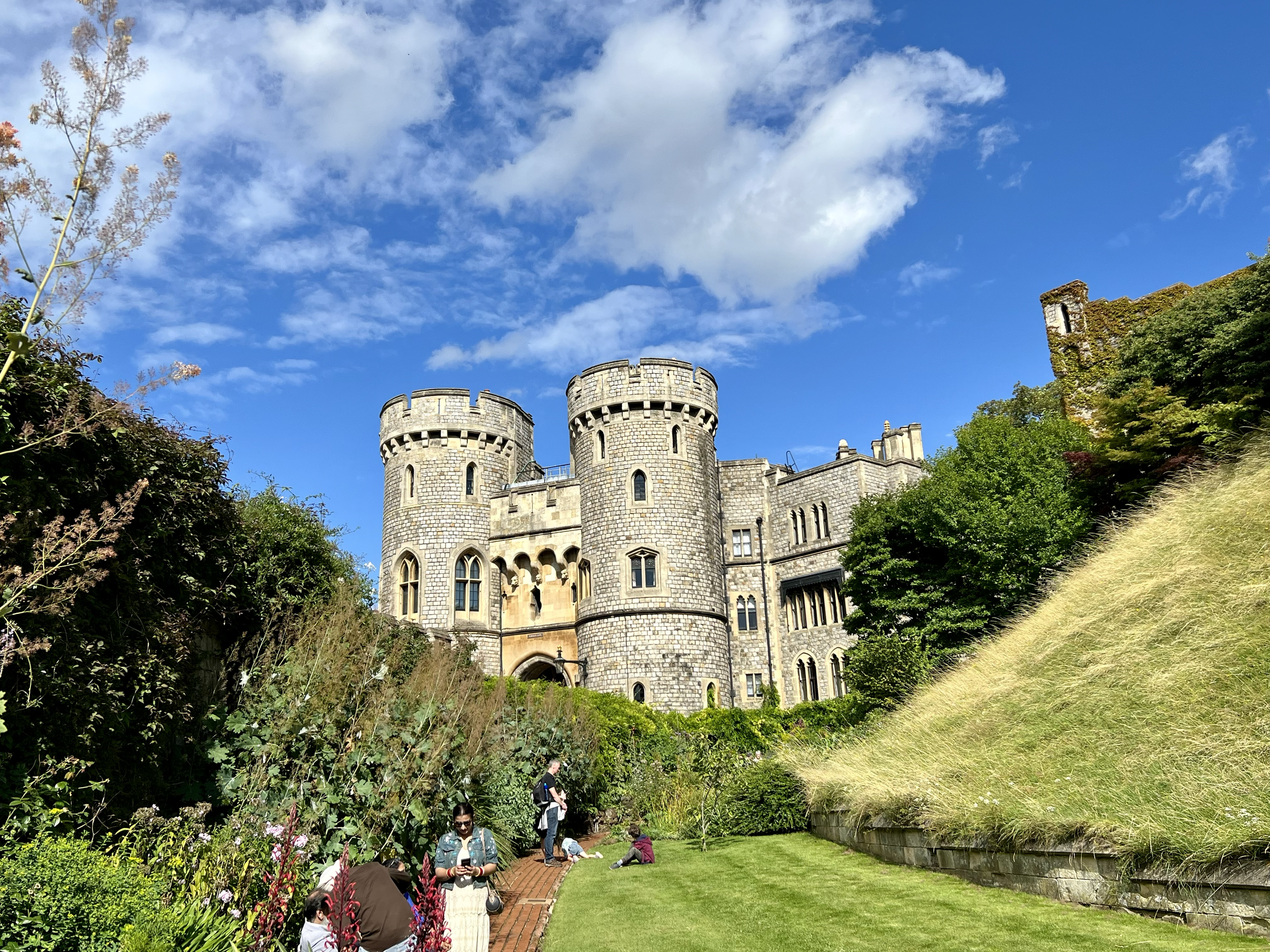 A castle with rounded towers and stone walls, surrounded by lush greenery and a garden, under a blue sky with scattered clouds.