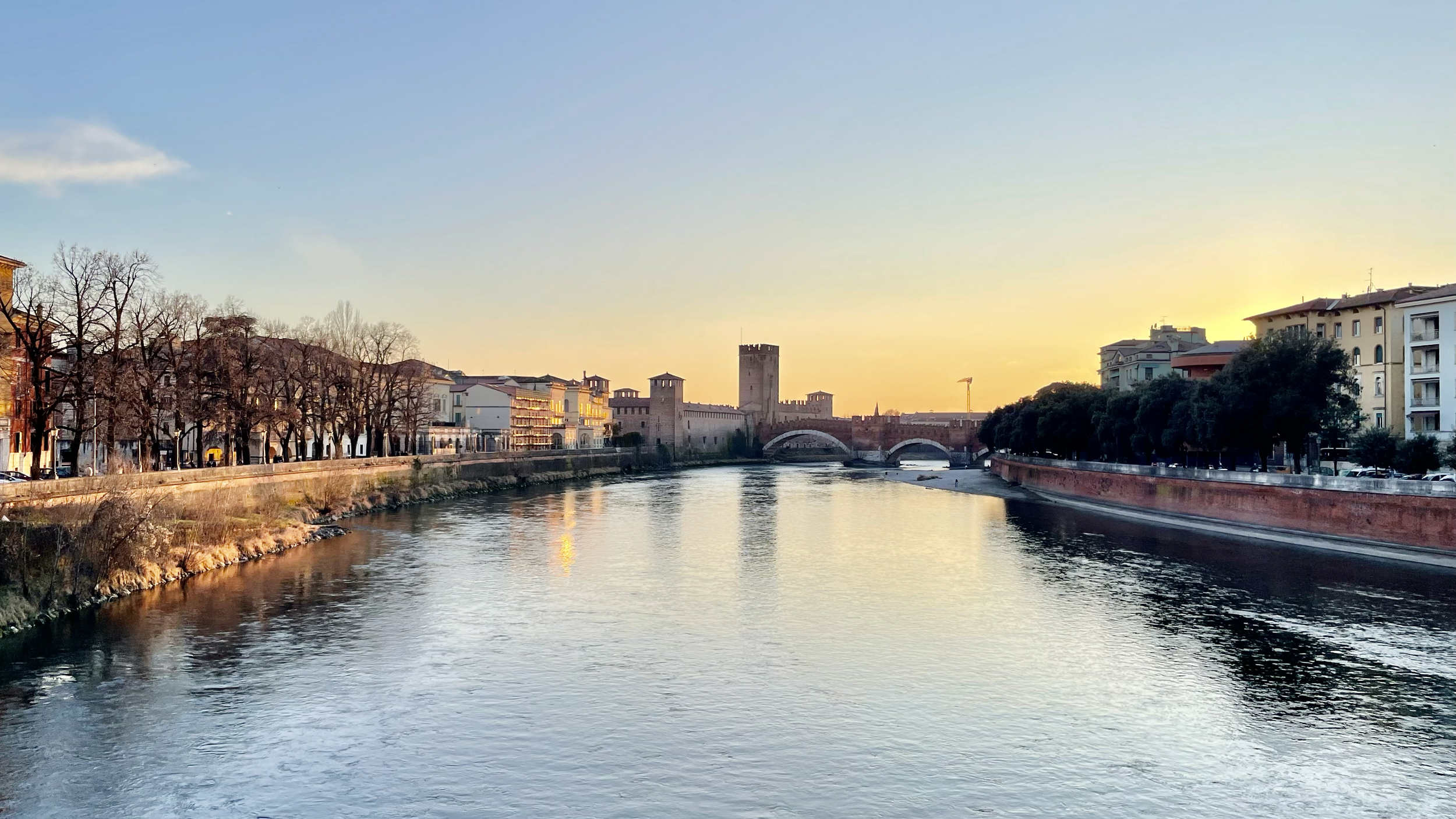 Sunset over a river with historic buildings and a bridge in the background, trees lining the riverbanks.