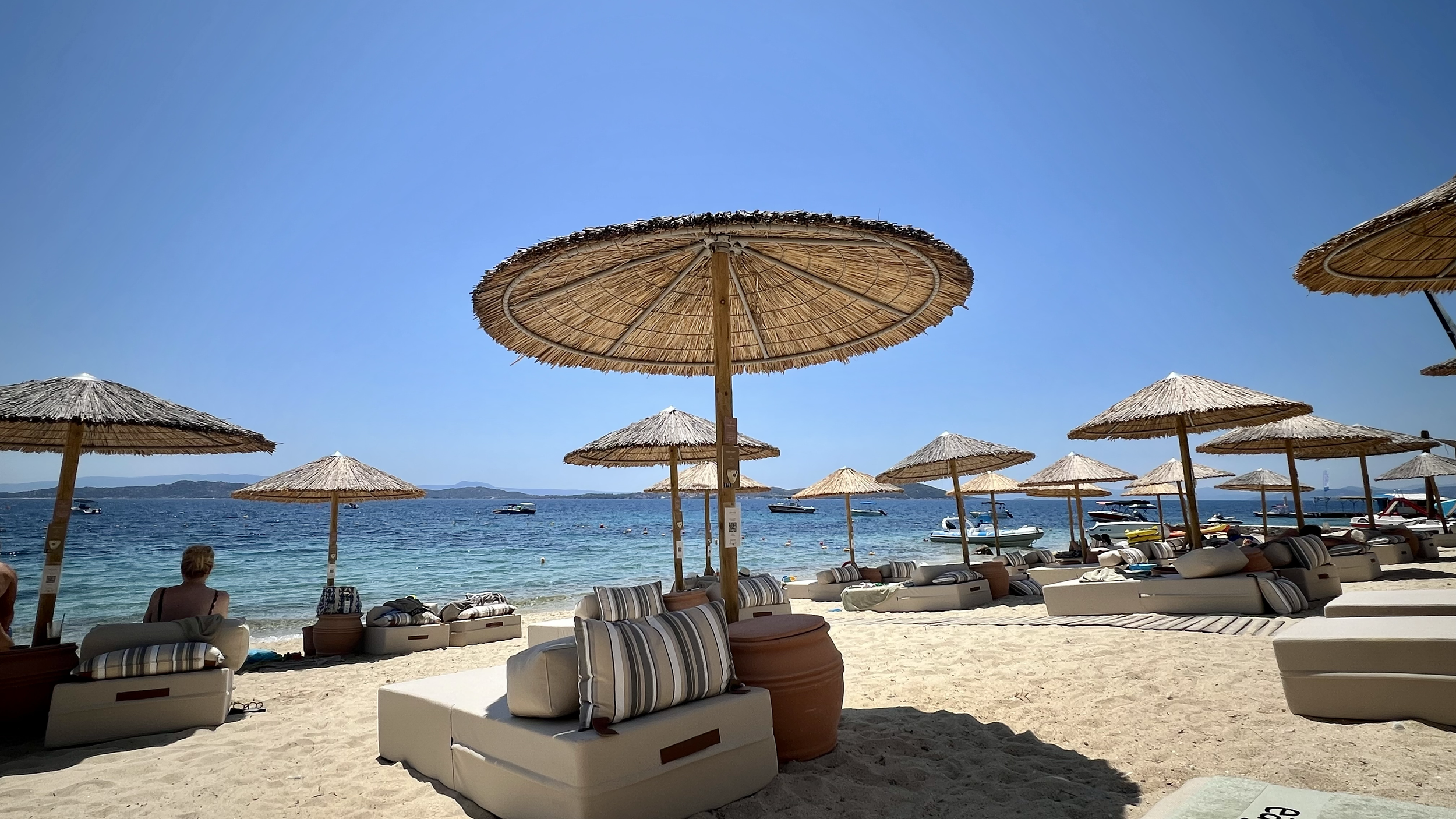 Beach scene with straw umbrellas, lounge chairs, and people relaxing by the ocean under a clear blue sky, with boats on the water and distant islands in the background.