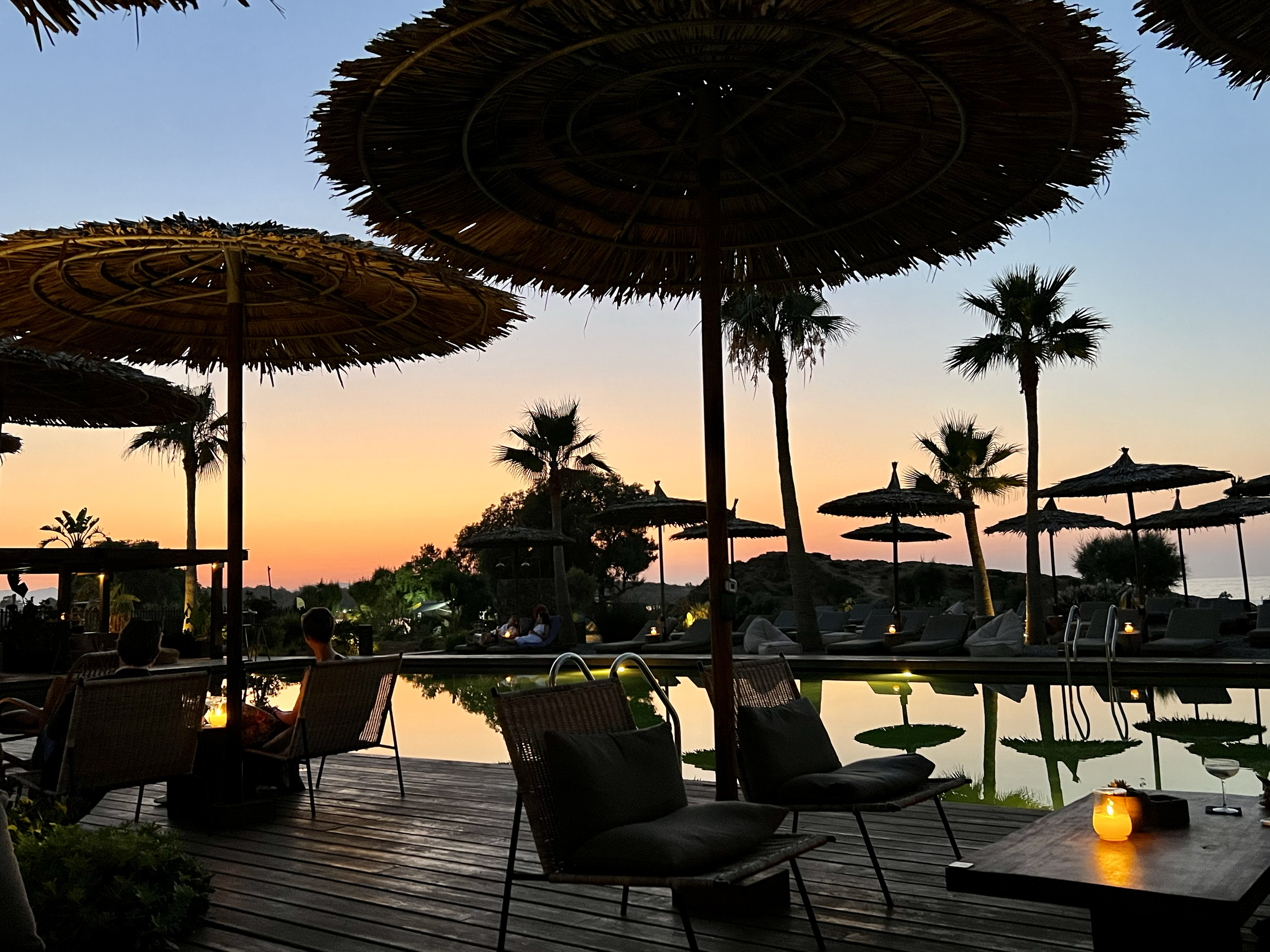 Sunset scene at a poolside bar with thatched umbrellas, lounge chairs, and palm trees, illuminated by candles.