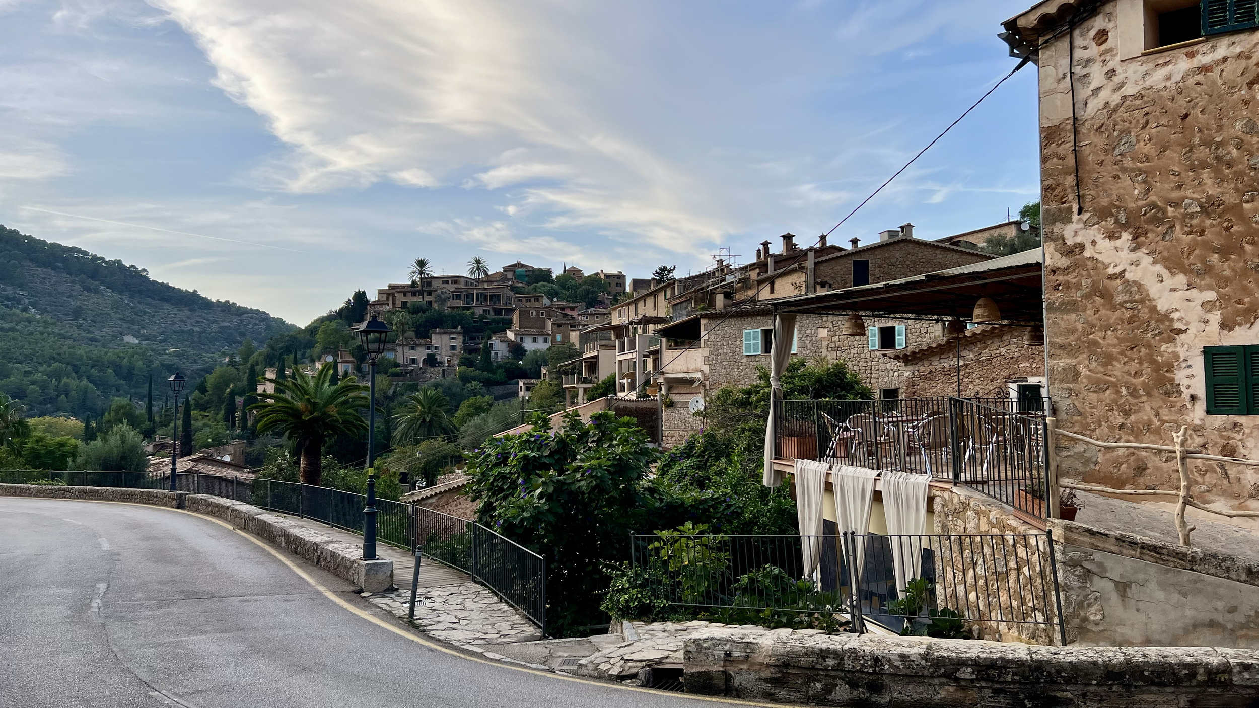 A scenic view of a hillside residential area with stone houses, palm trees, and lush greenery, taken from a winding paved road with streetlights and a metal railing.