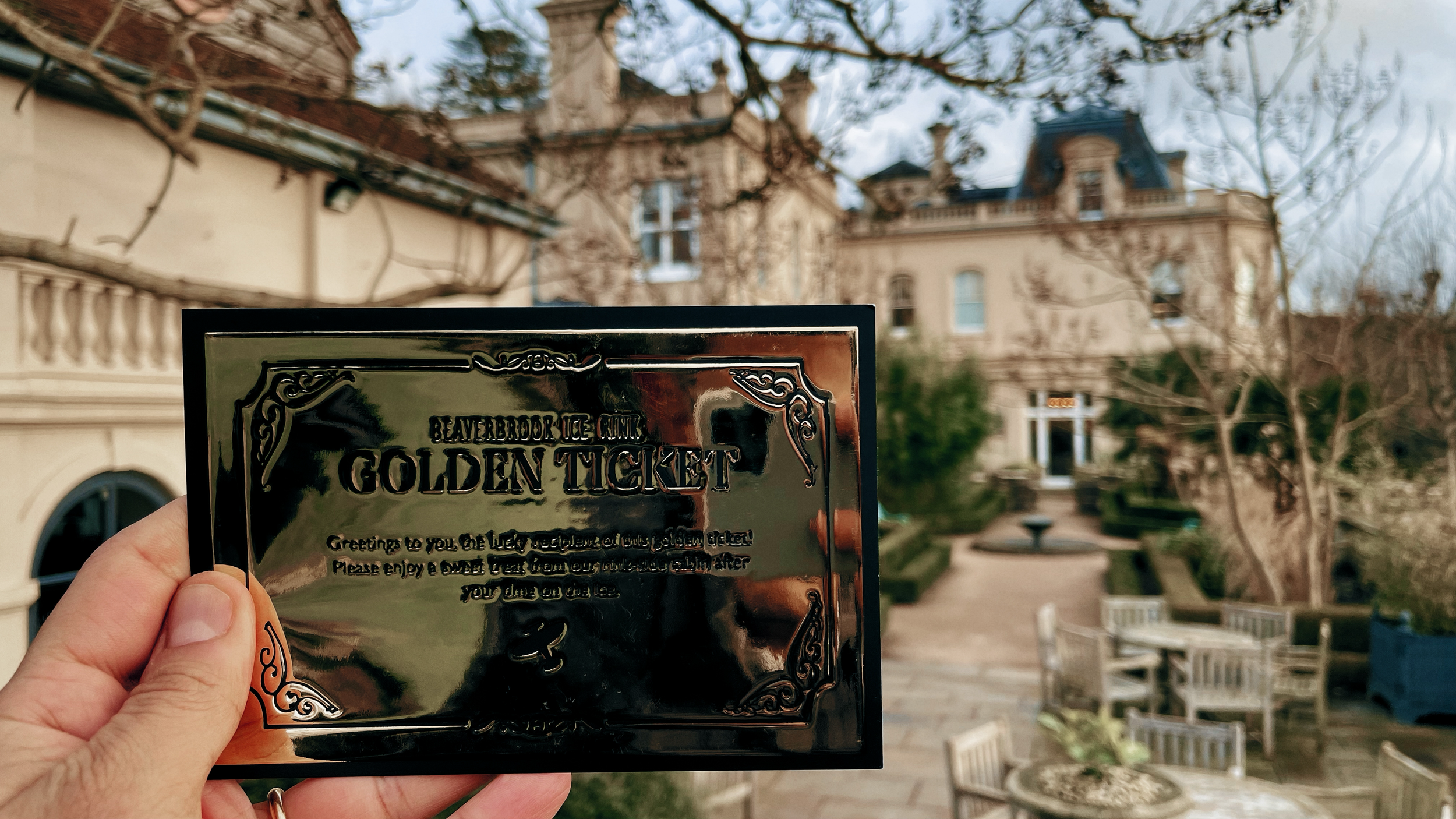 A hand holding a shiny black ticket for the Beaverbrook Ice Club, with a luxurious large house and a patio with chairs and tables in the background.