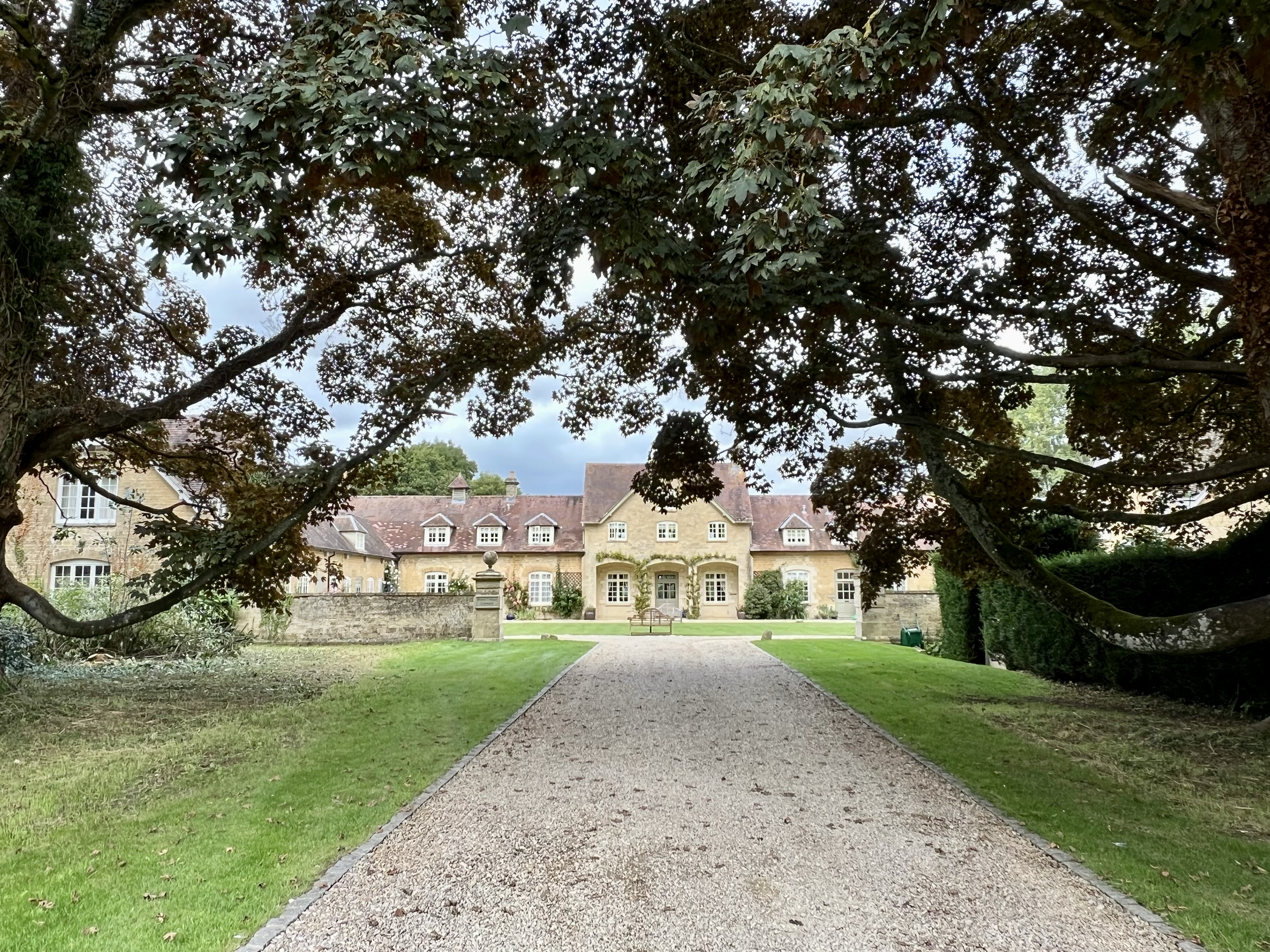 A large mansion with a stone pathway leading up to it, framed by overhanging tree branches. The house has multiple windows, a central entrance, and a neatly maintained lawn.