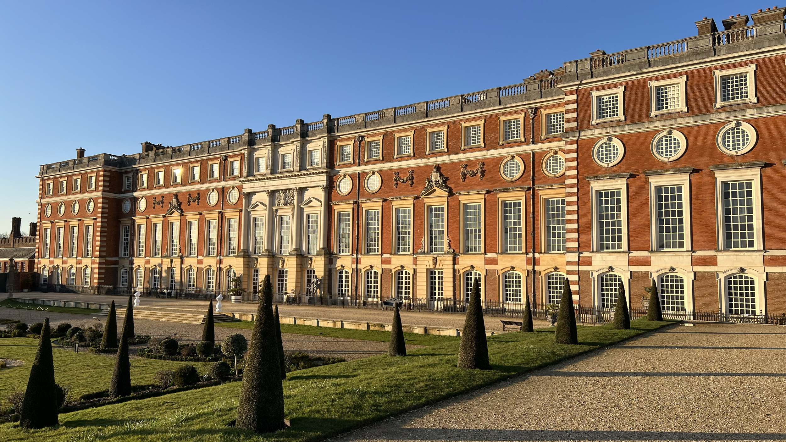 A grand historic mansion with a red brick facade, tall windows, decorative stone accents, and a landscaped garden with trimmed bushes and conical topiary under a clear blue sky.