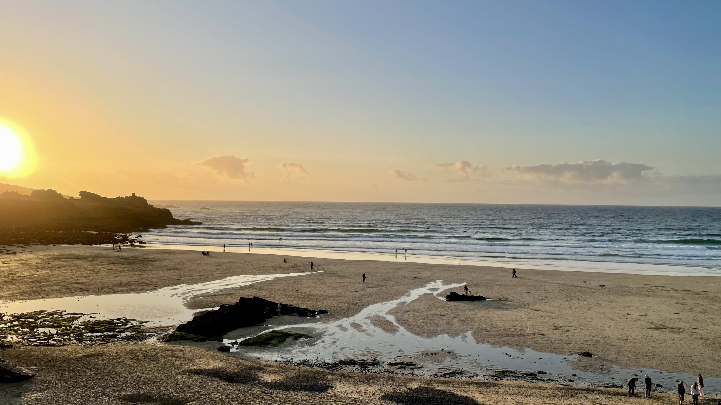 Sunset over a sandy beach with scattered rocks, small pools of water, and people walking or sitting along the shoreline with a rocky cliff on the left and the ocean stretching to the horizon.