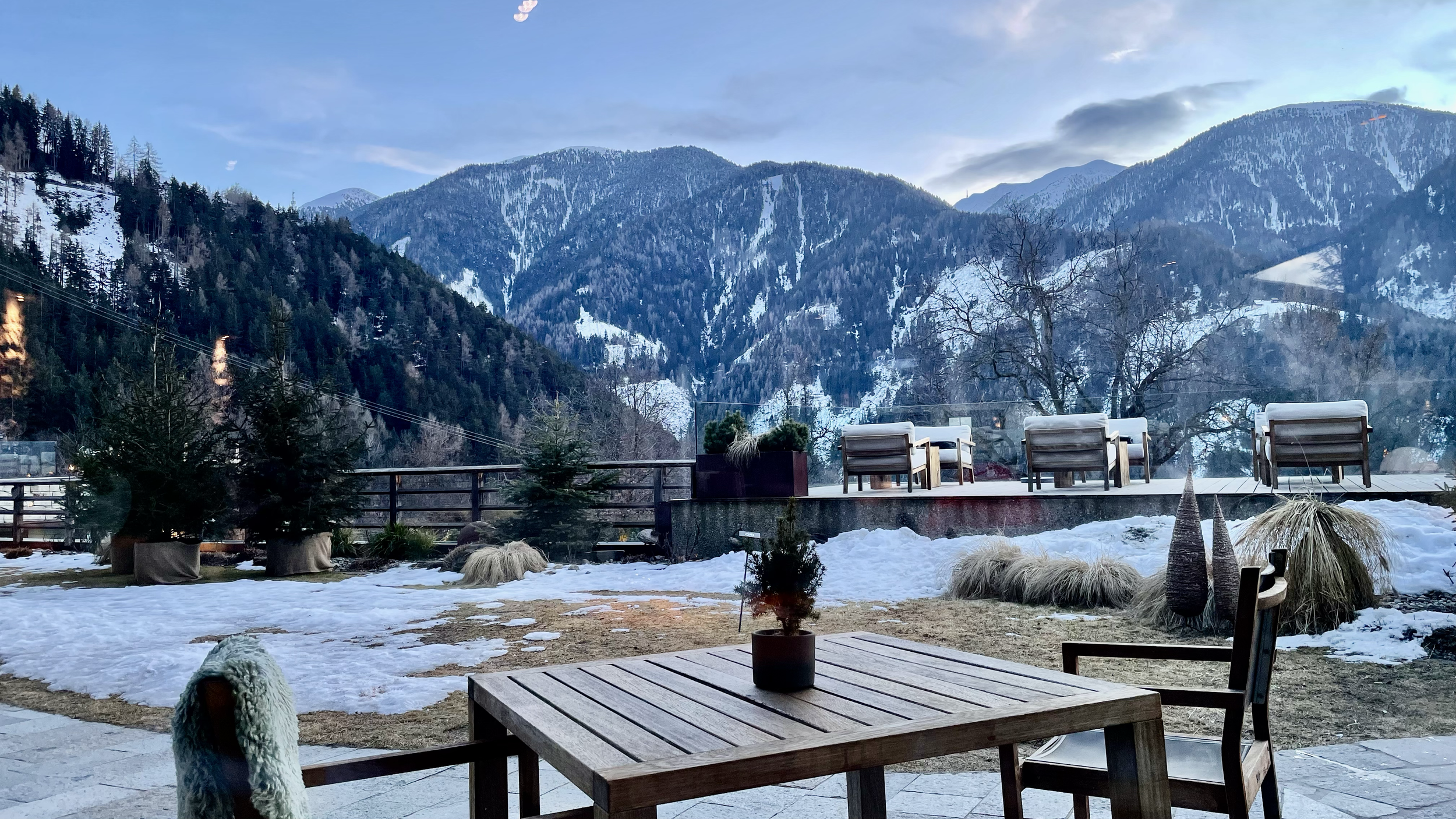 Outdoor patio with wooden table and chairs, potted plants, snow-covered ground, mountains in the background, and some trees.