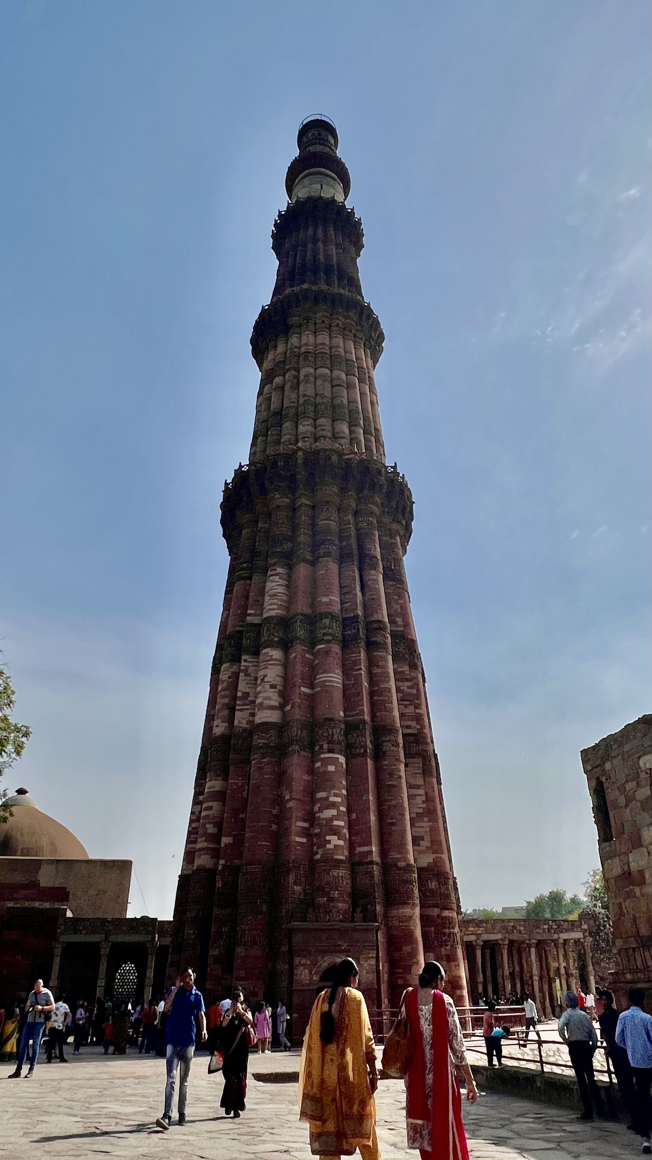 People walking near a tall, ancient brick minaret against a clear blue sky, with historic buildings and ruins around.