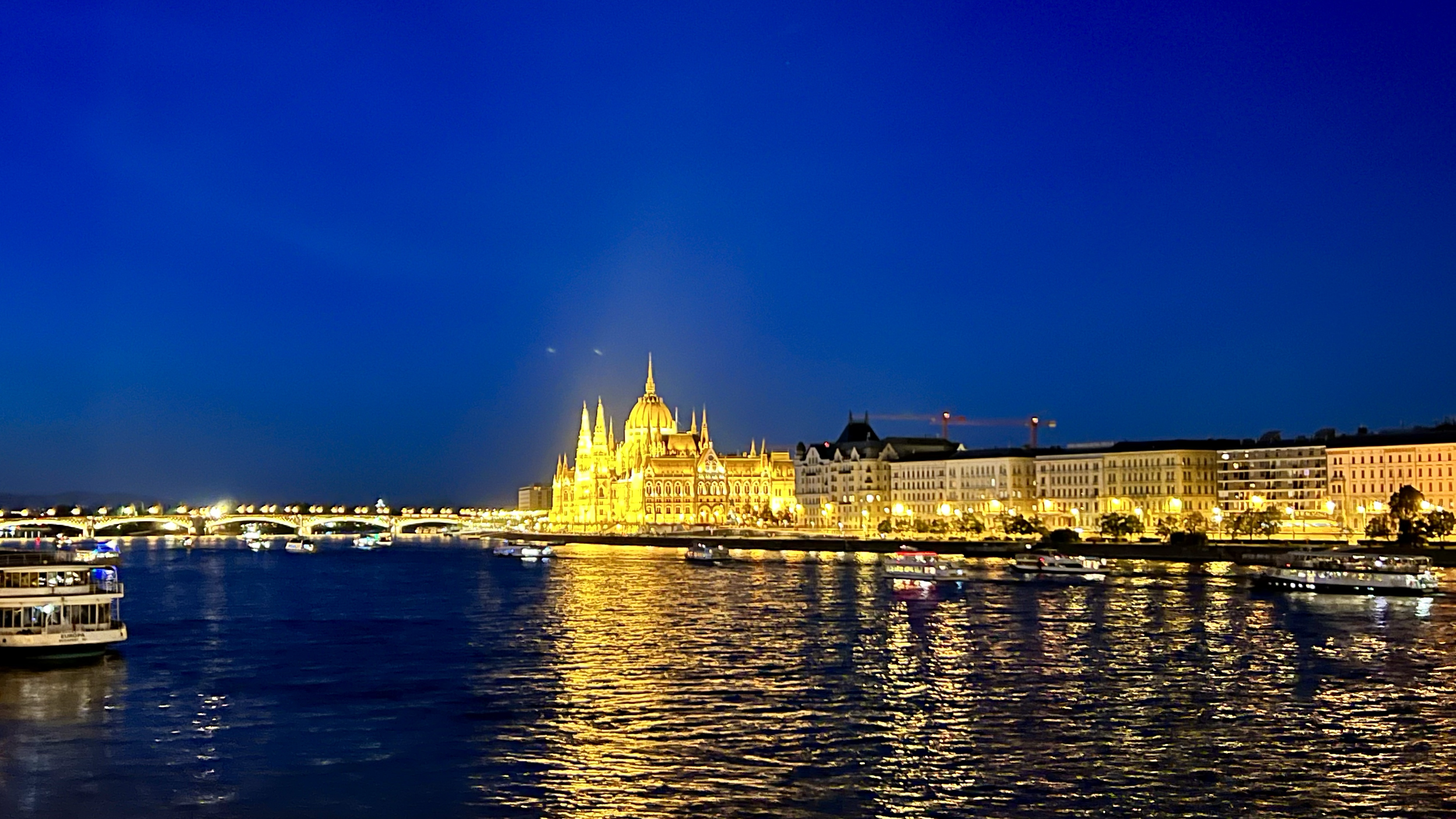 Nighttime view of a cityscape with illuminated historic buildings and a river with boats reflecting the city lights.