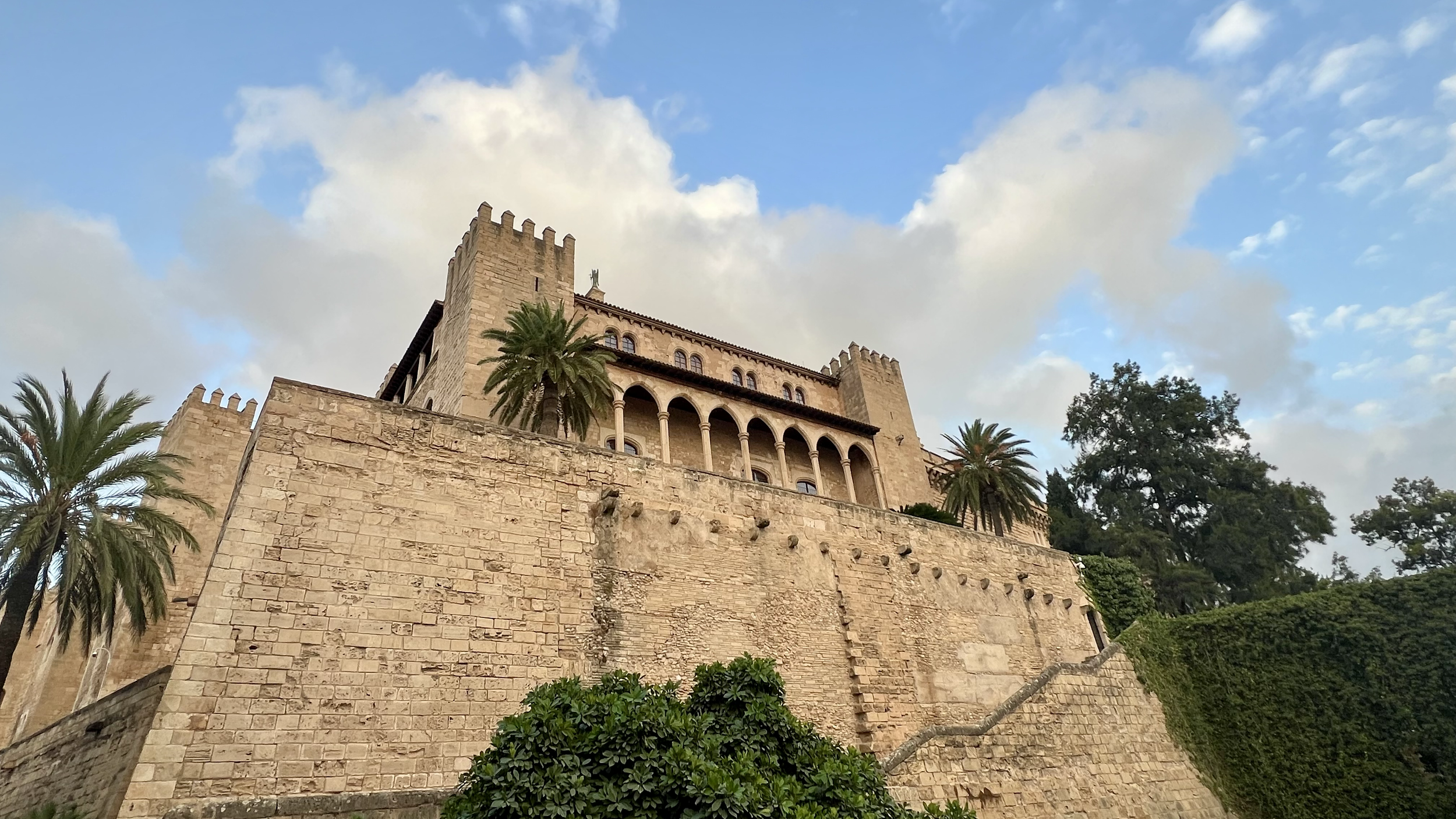 A historic castle with beige stone walls, palm trees, and a partly cloudy sky.