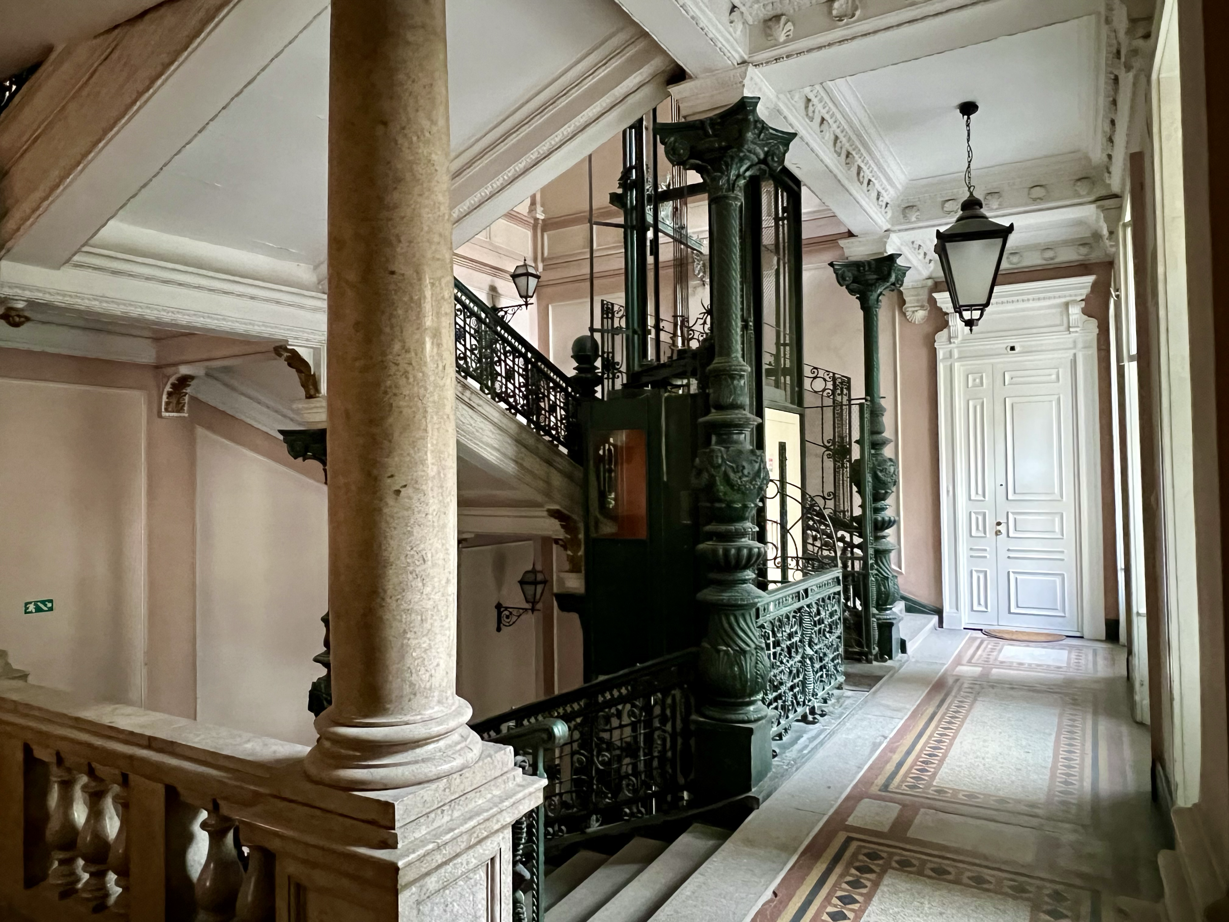 Interior view of an elegant building with ornate columns, decorative moldings, a staircase with intricate railings, and vintage lighting fixtures.