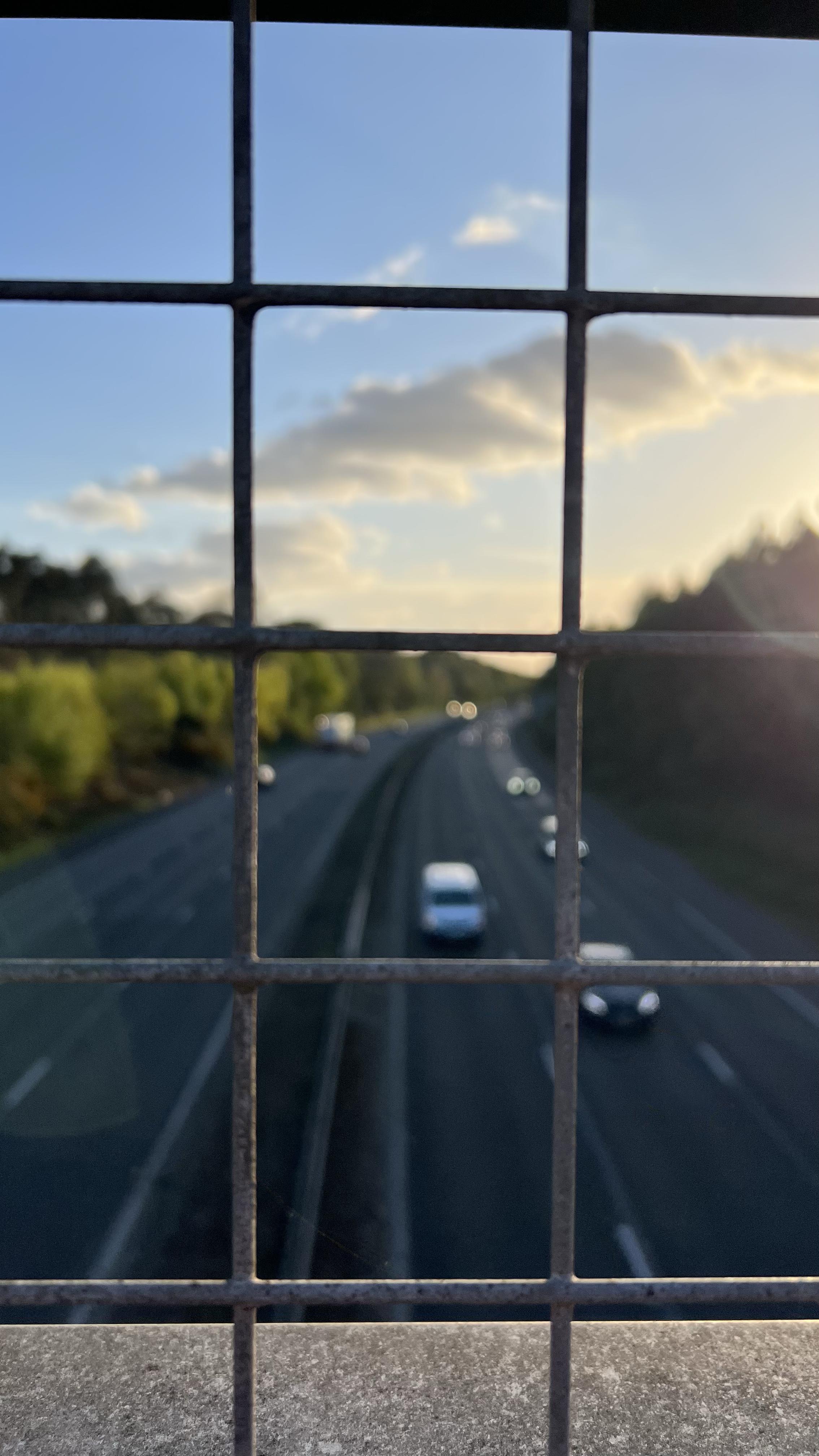 View of a highway with cars passing through, seen through a metal grid fence at sunset with clouds in the sky.